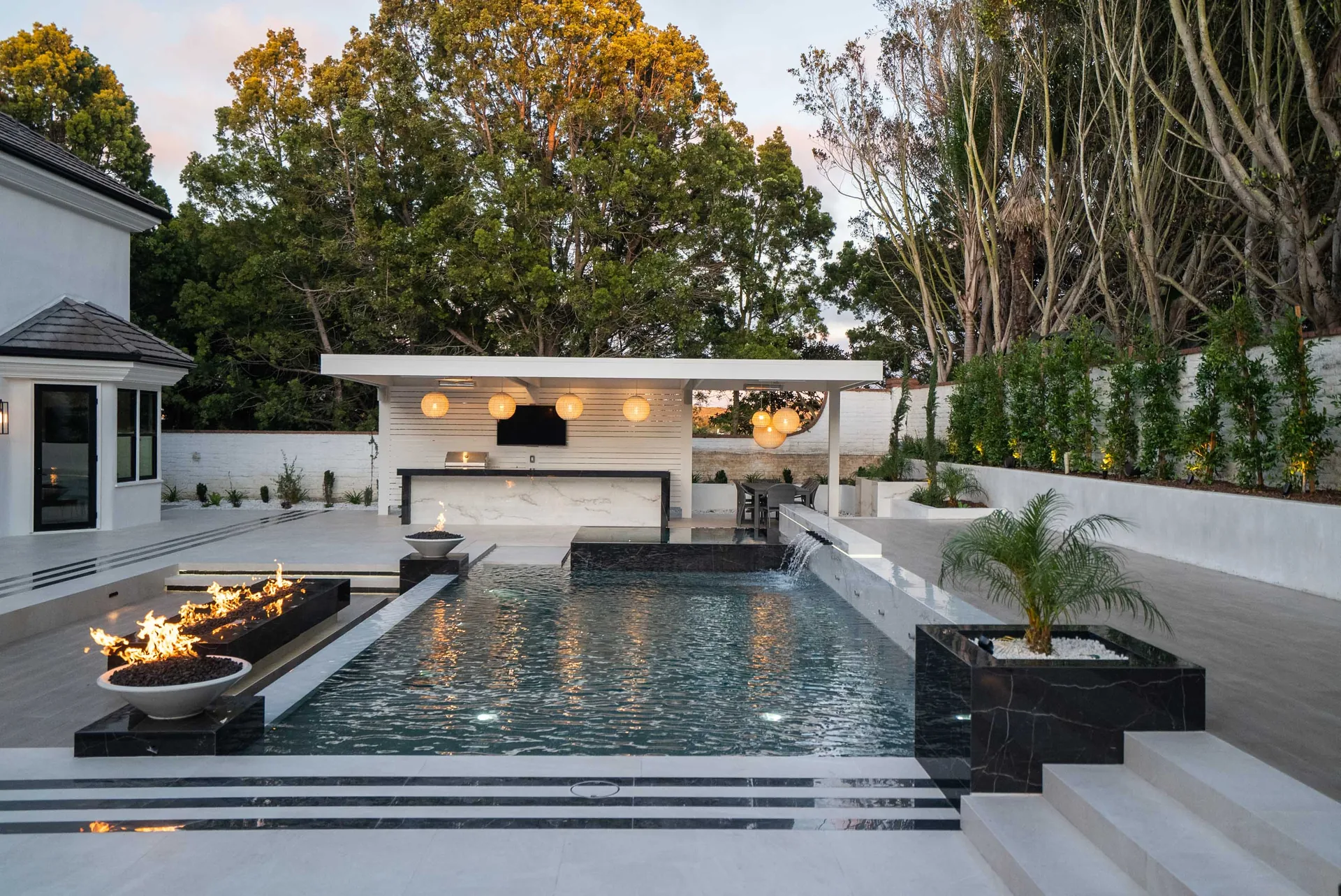 The pool running toward the covered kitchen at dusk, a long fire trough burning along one side and mature trees standing behind.
