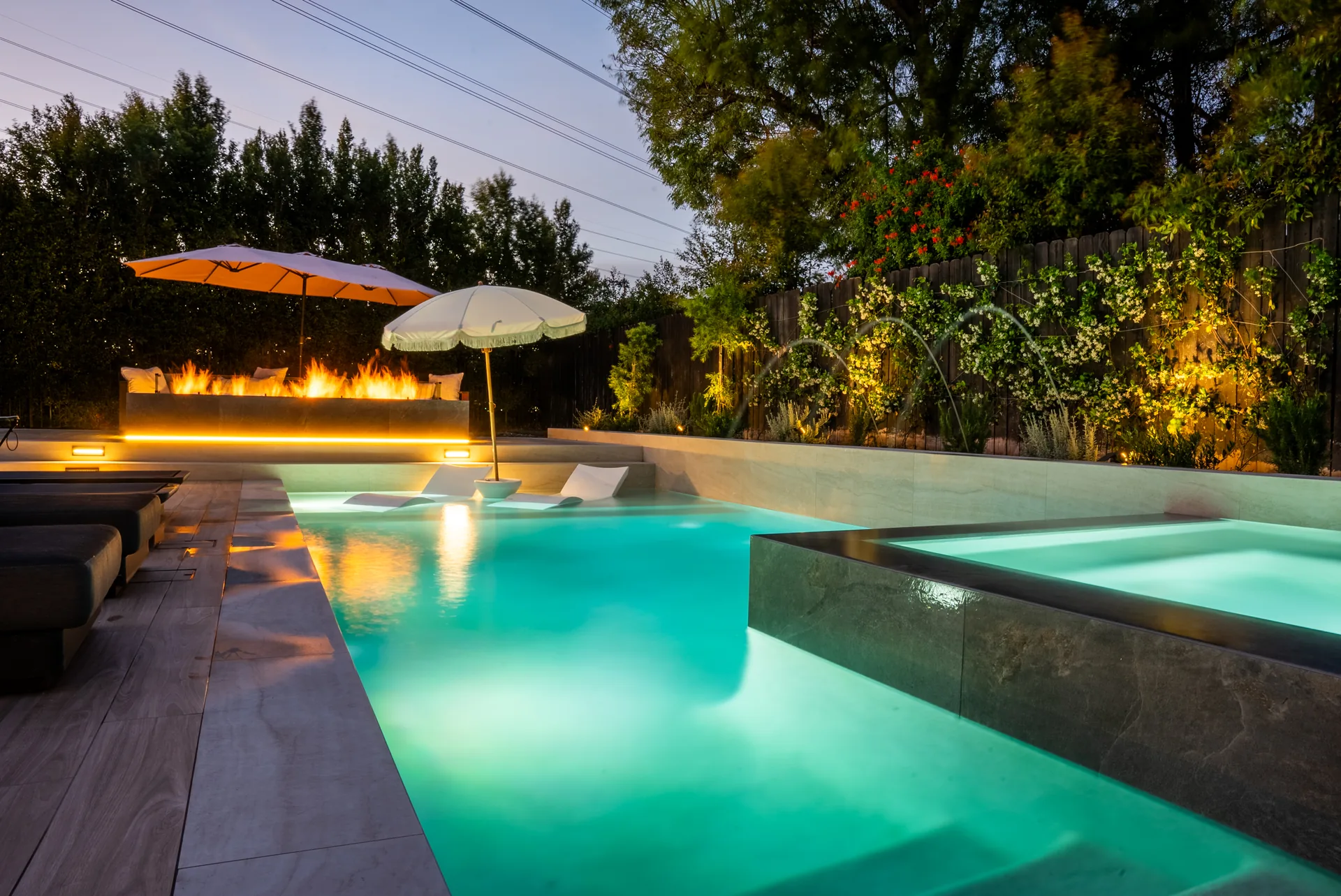 The pool lit turquoise at dusk with a long fire feature burning along the raised wall behind, umbrellas and loungers on the near deck.