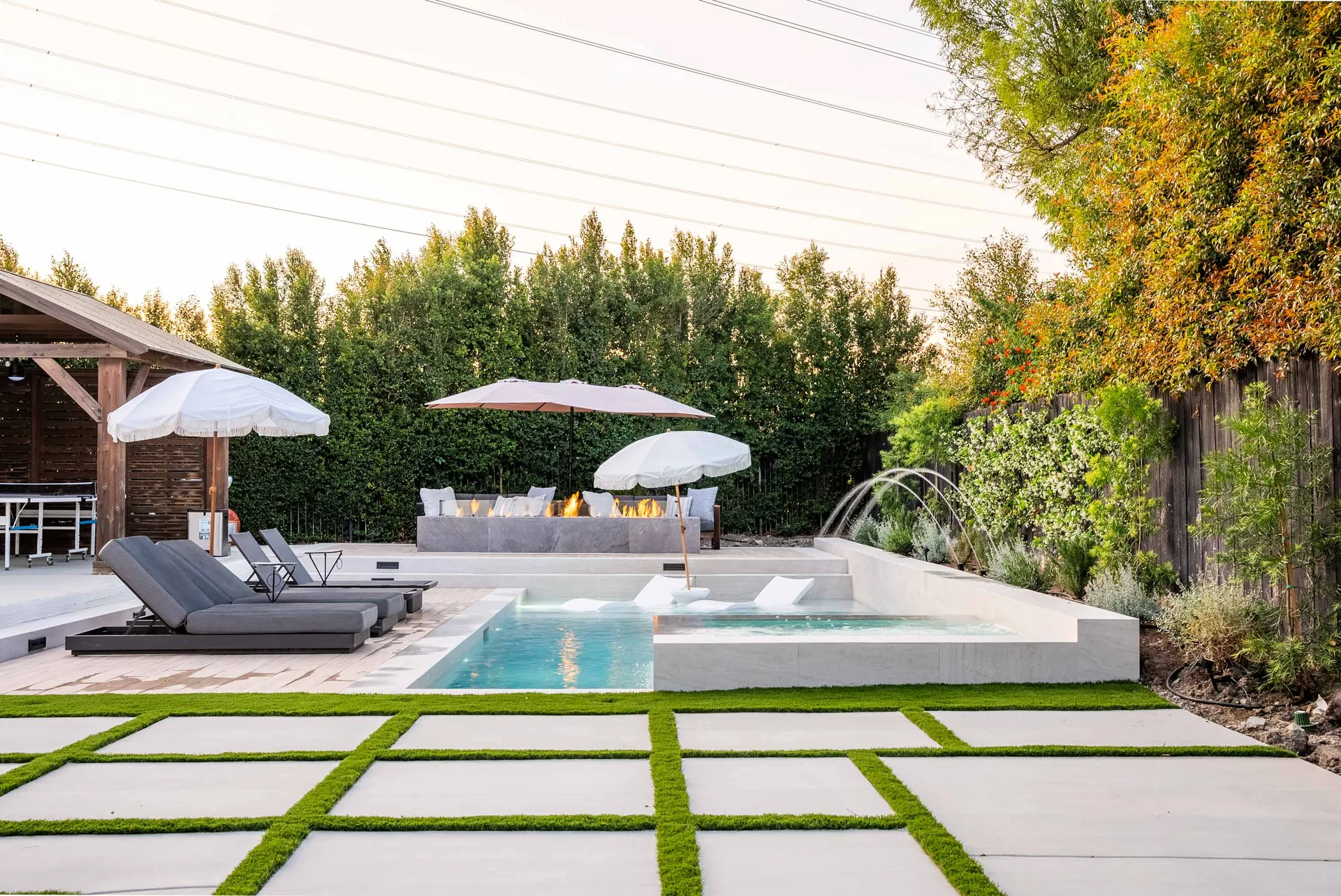 The yard in daylight, with umbrellas over loungers, deck jets arcing into the pool and paving set into strips of turf.