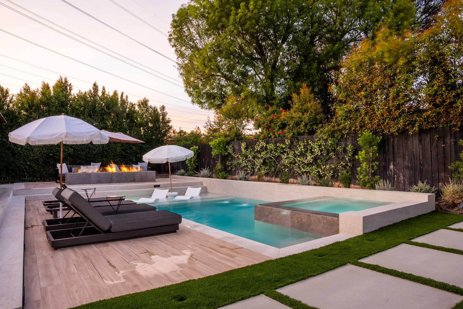 The pool from the deck with dark loungers under a parasol, a raised spa at the far end and turf running alongside.