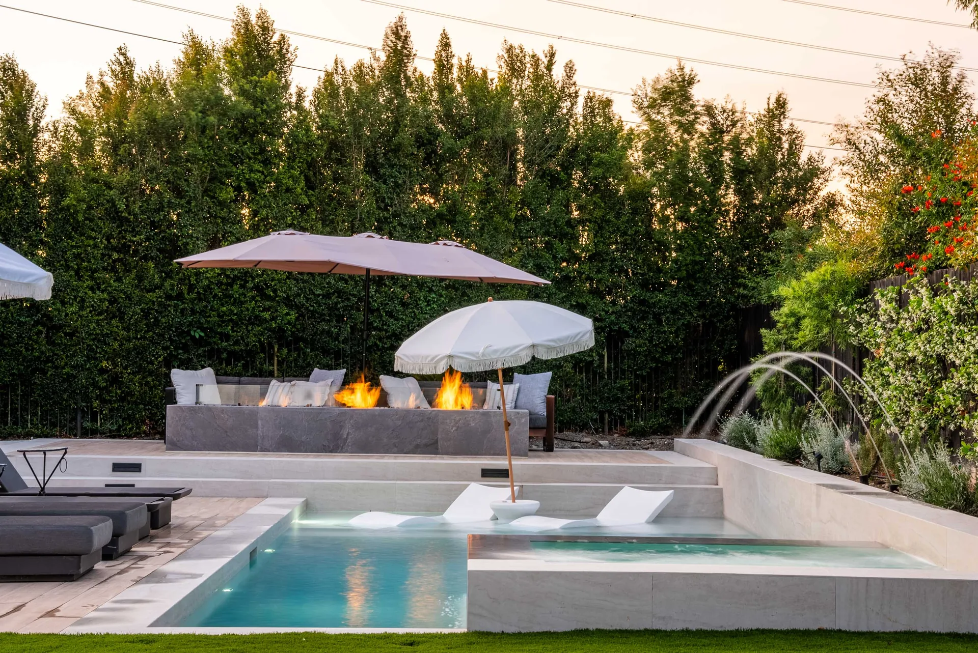 A fire feature burning on the raised wall beside the pool, parasols above and in-water loungers on the tanning shelf.