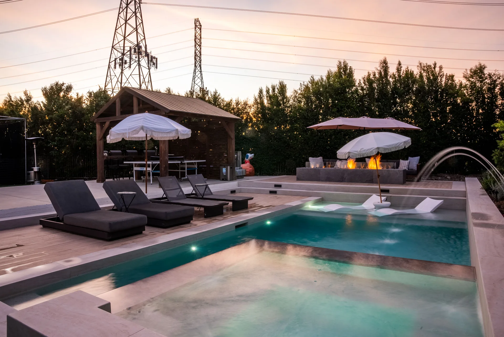 The yard at dusk from the far corner, umbrellas and loungers along the pool with a timber pavilion at the end.