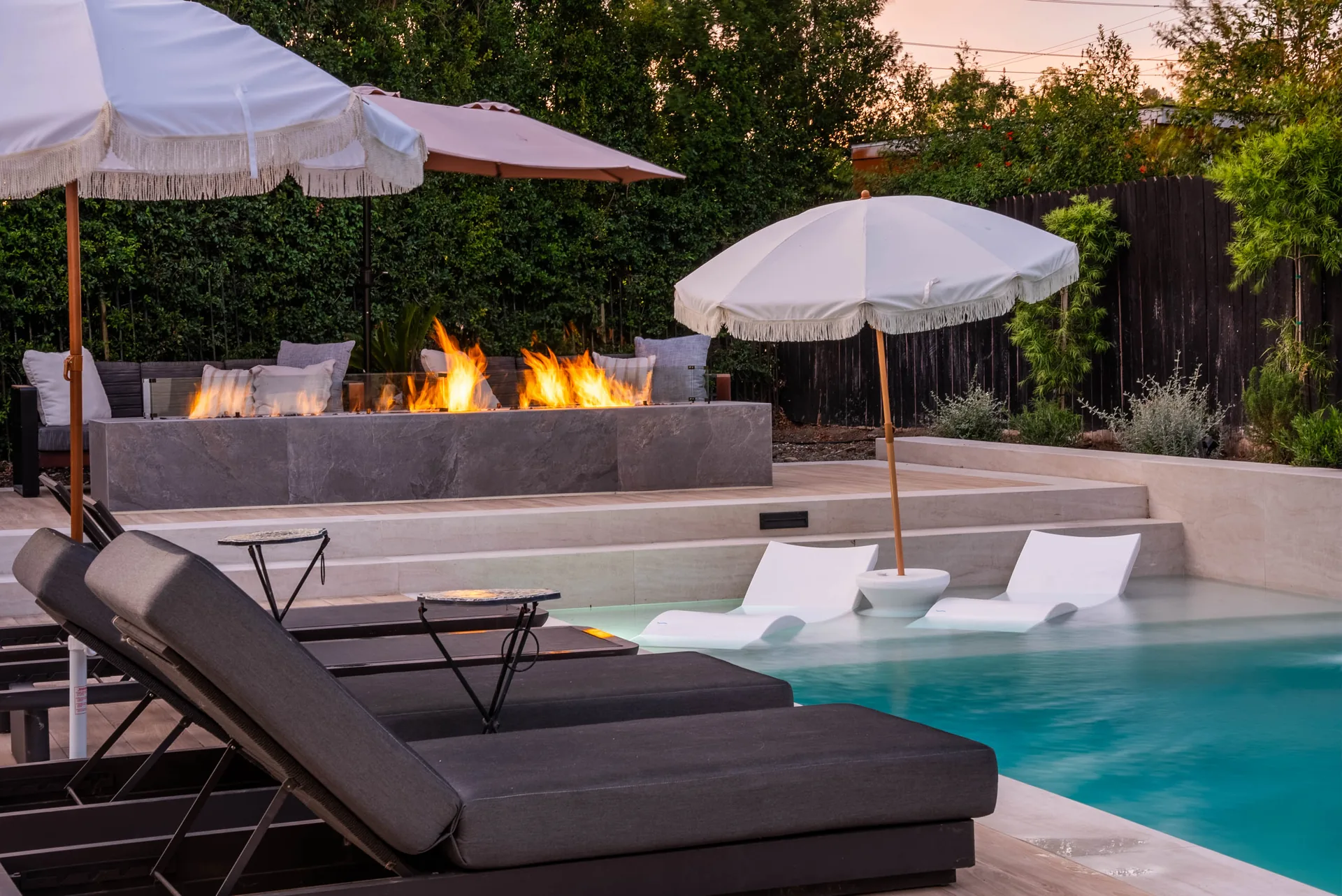 Two fringed parasols over dark loungers, the fire feature burning on the wall behind and white loungers standing in the shallow water.