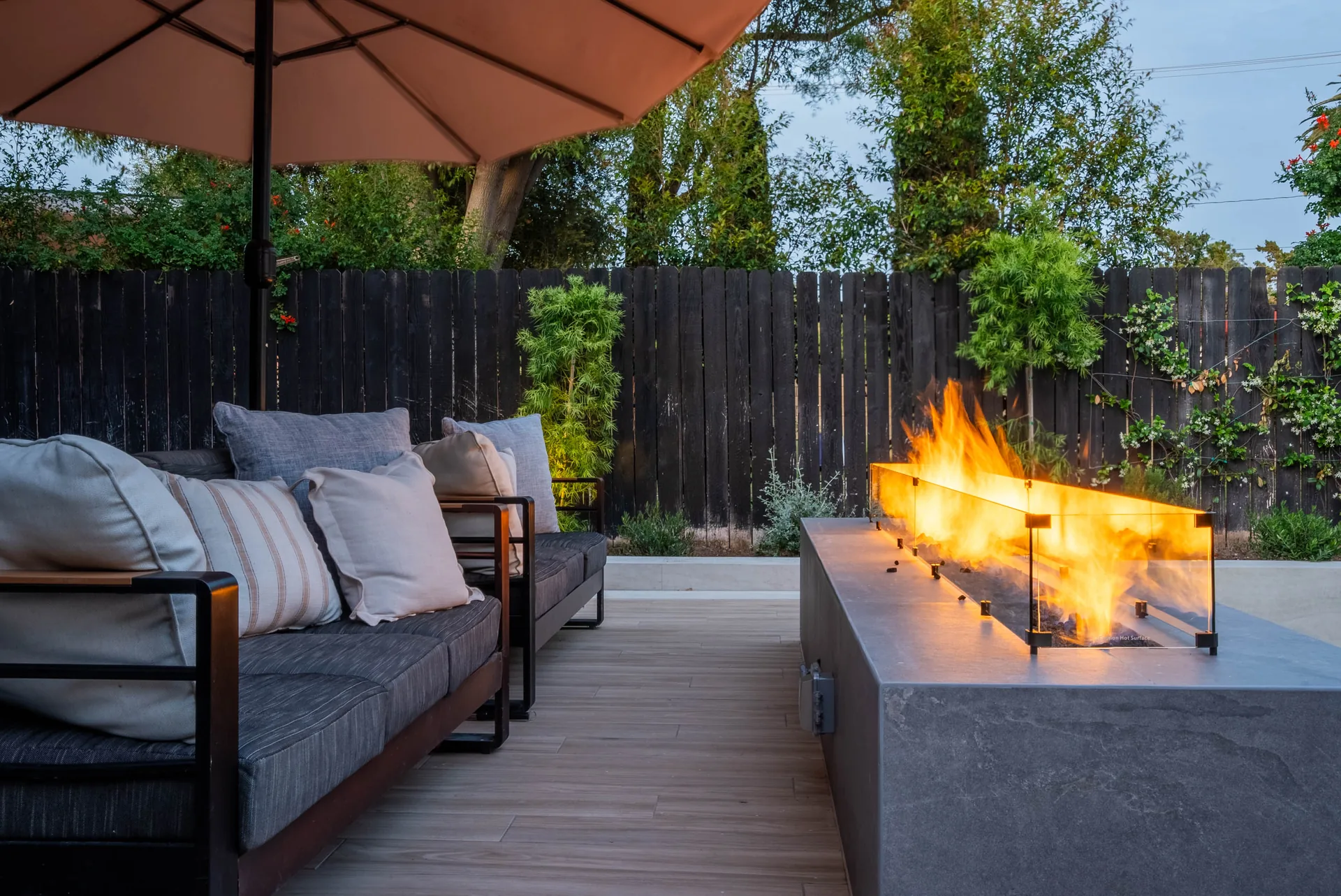 A cushioned sofa facing a long concrete fire trough burning on the deck, dark fence and planting behind.