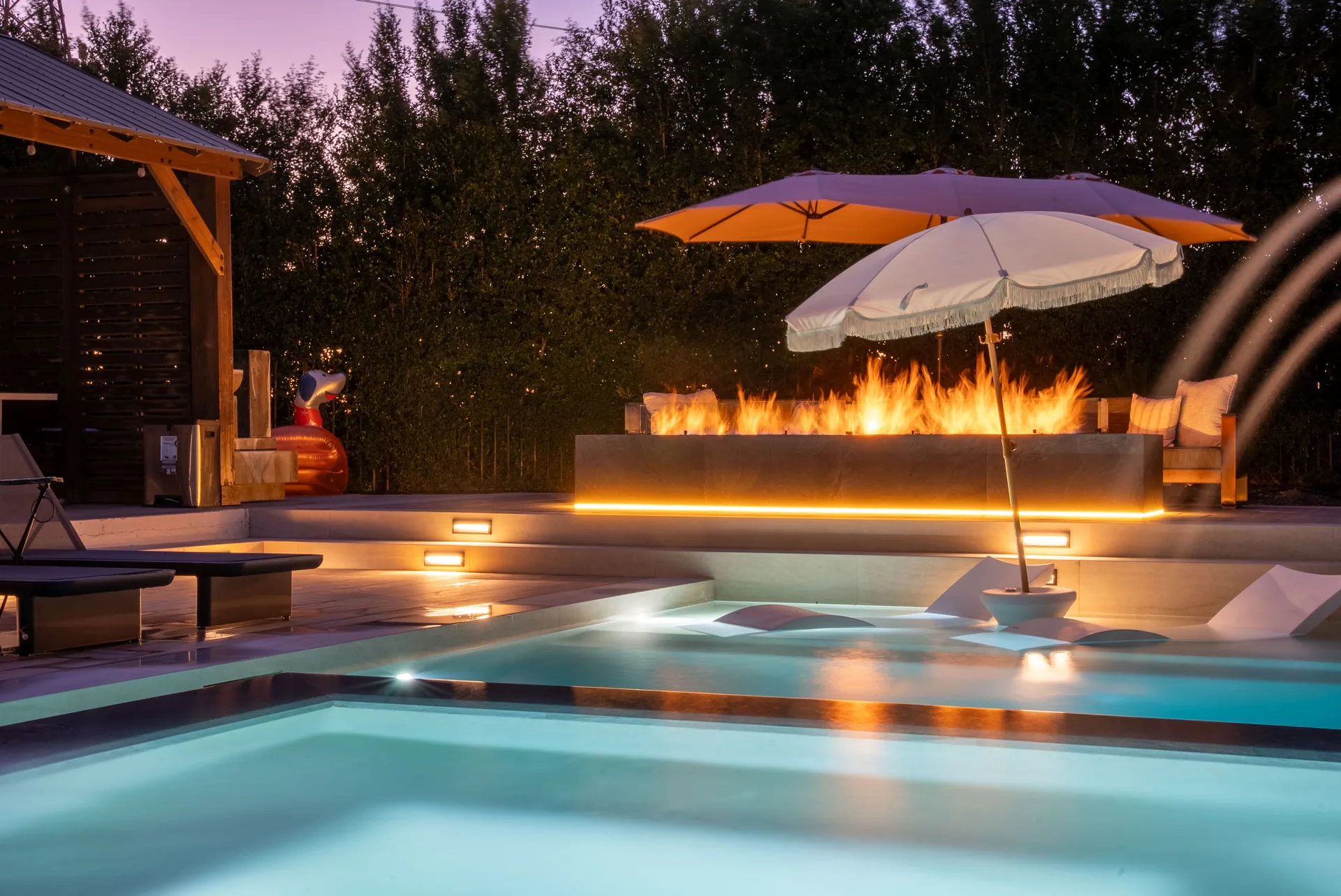 The pool and spa after dark under a violet sky, deck jets arcing and the fire feature burning behind.