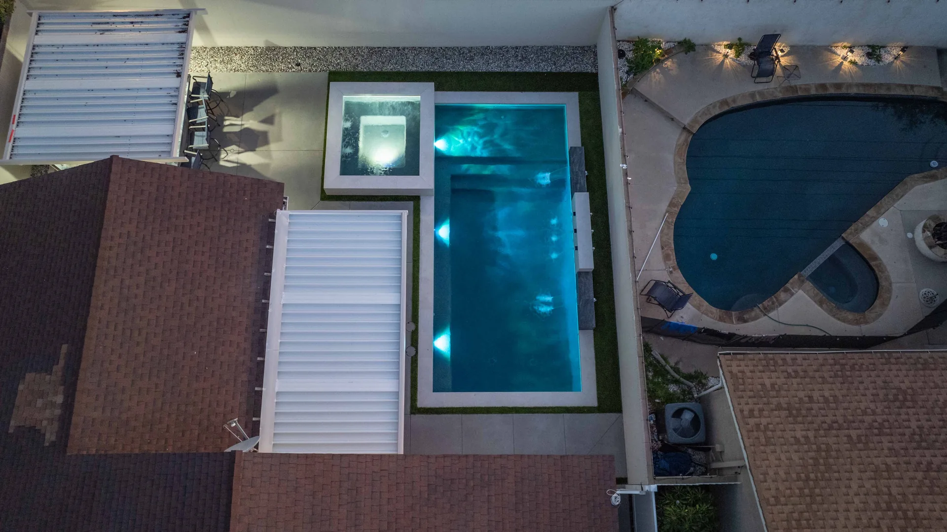 The yard from overhead at dusk, the new pool lit turquoise beside the older kidney-shaped pool it replaced.