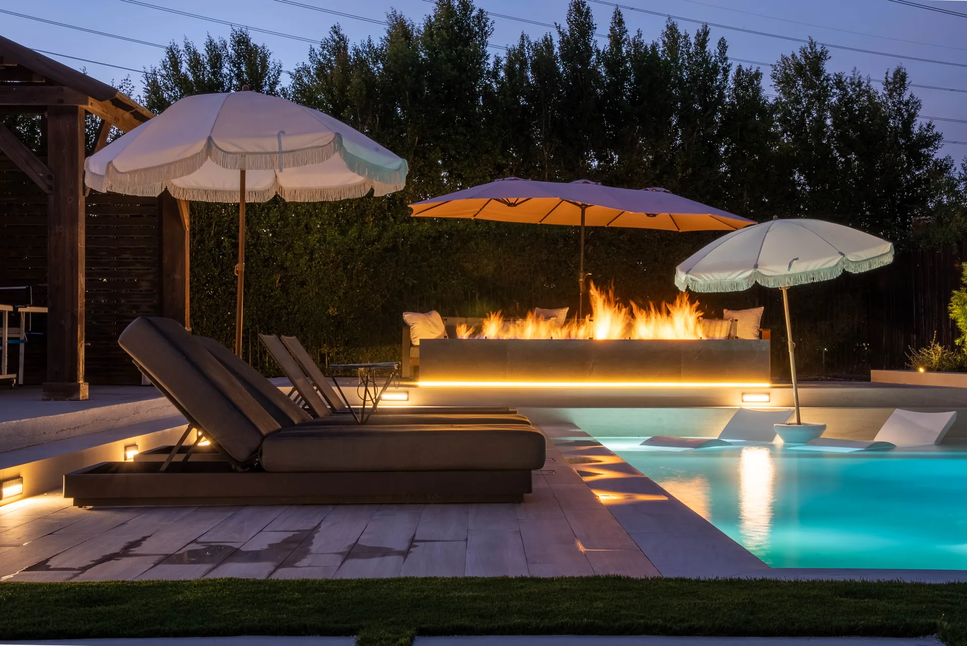 Loungers and fringed parasols along the deck at night, the fire wall lit behind and the pool glowing beside them.