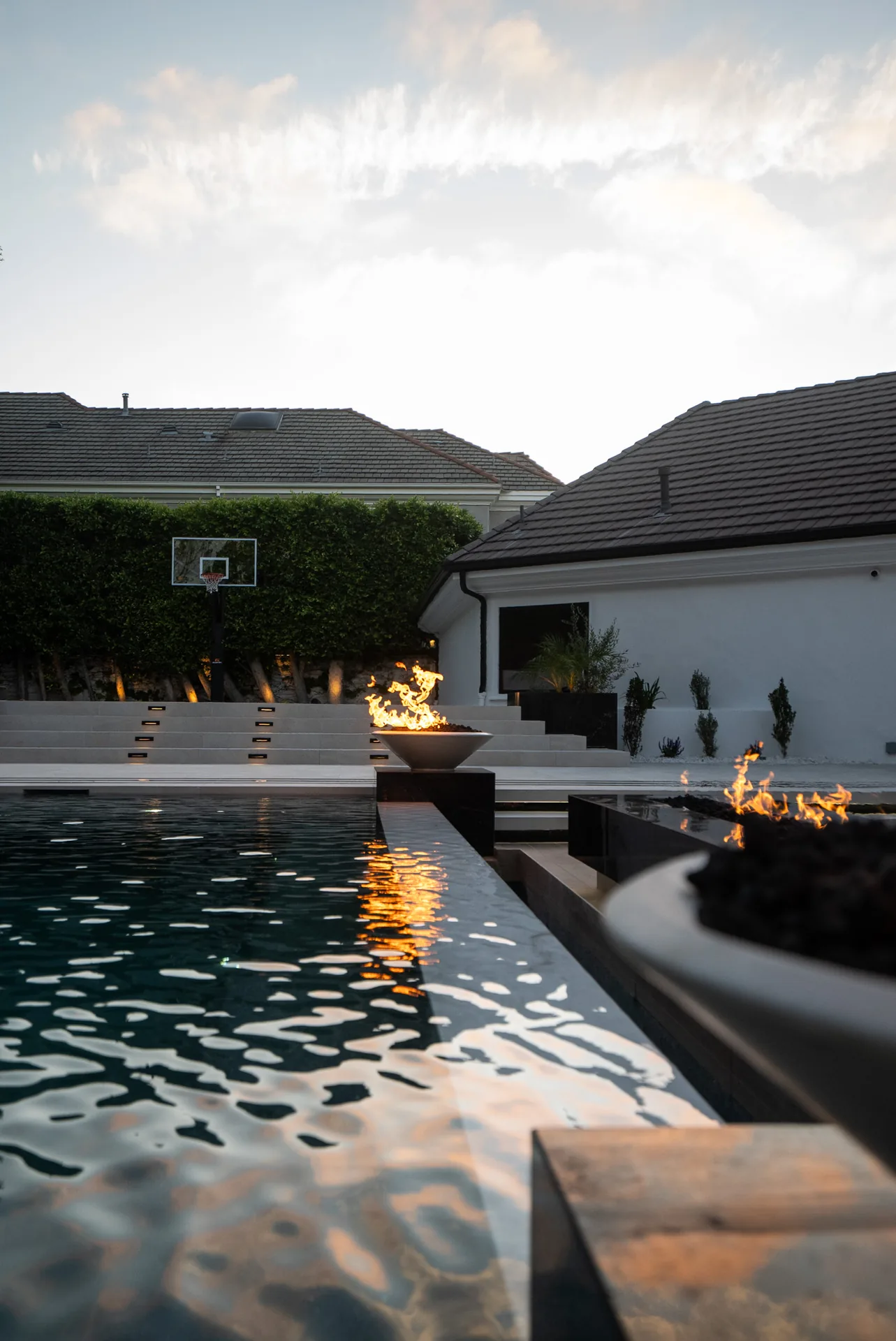 A black-tiled pool holding a still, dark surface, with a low fire bowl burning at its edge and a single-storey house beyond.