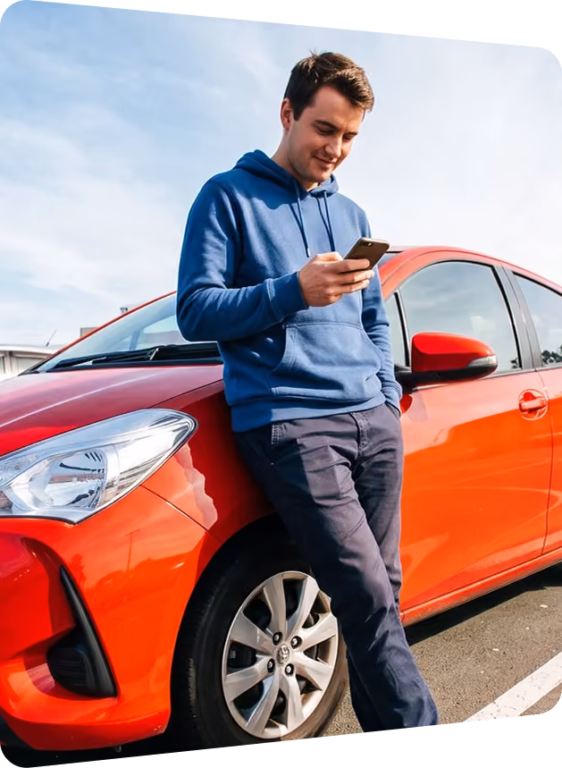 Man in a blue hoodie leaning on a red hatchback while checking his phone