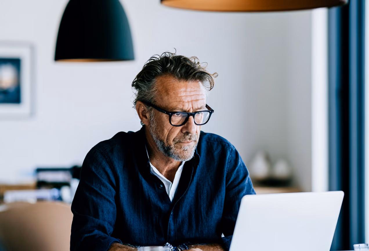 Homme d'âge moyen portant des lunettes, assis à une table, concentré sur un ordinateur portable dans un intérieur lumineux et moderne.