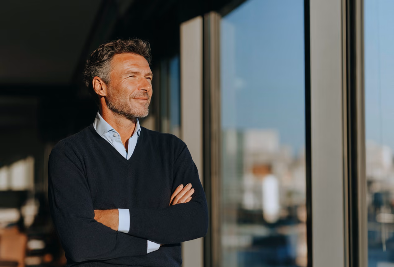 Homme d'affaires souriant en chemise blanche sous un pull noir, bras croisés, regardant par une grande fenêtre vitrée avec réflexion de la ville en arrière-plan.