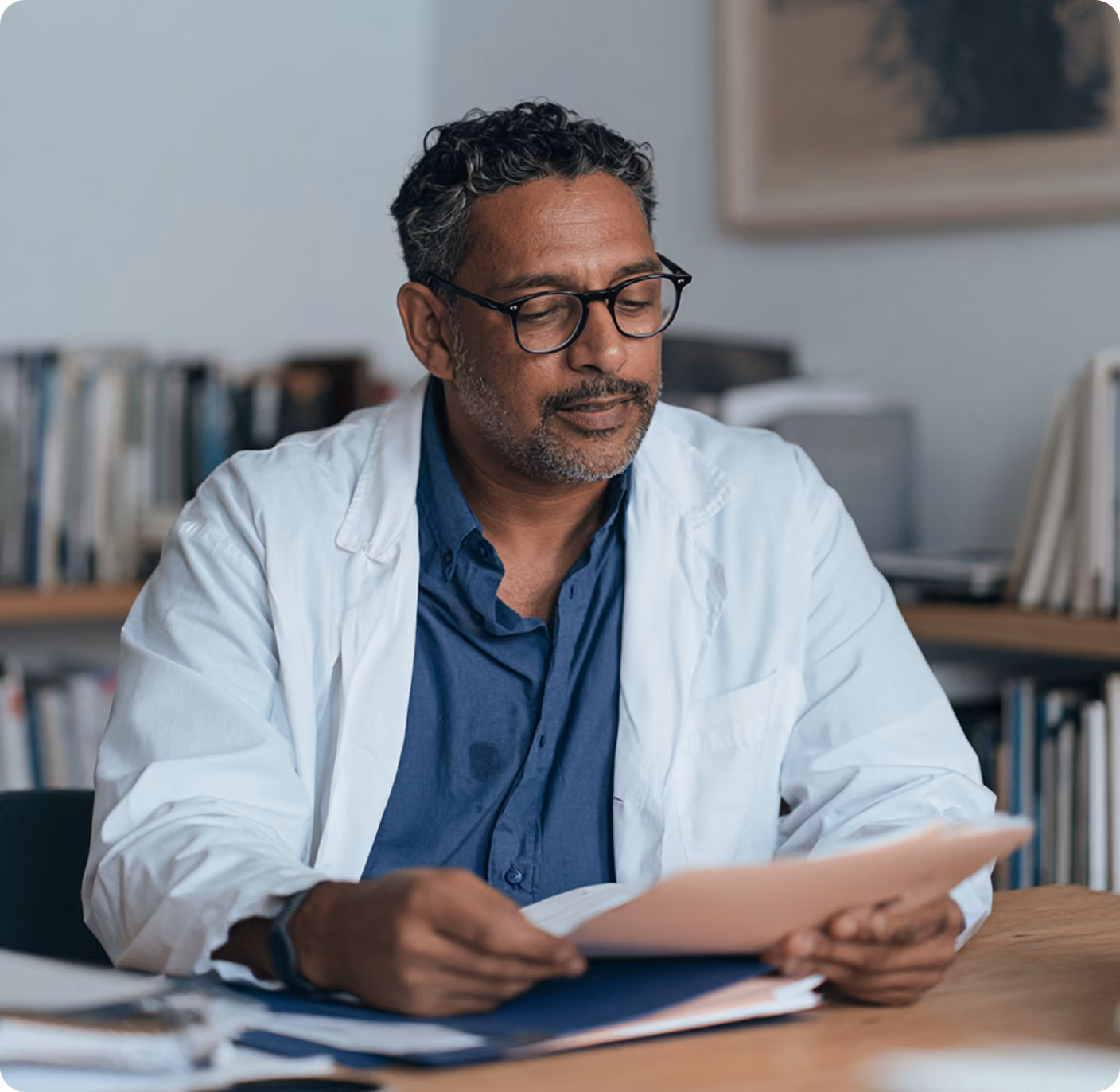 Médecin portant une blouse blanche et des lunettes lisant des dossiers assis à son bureau dans un cabinet médical avec des étagères de livres en arrière-plan.