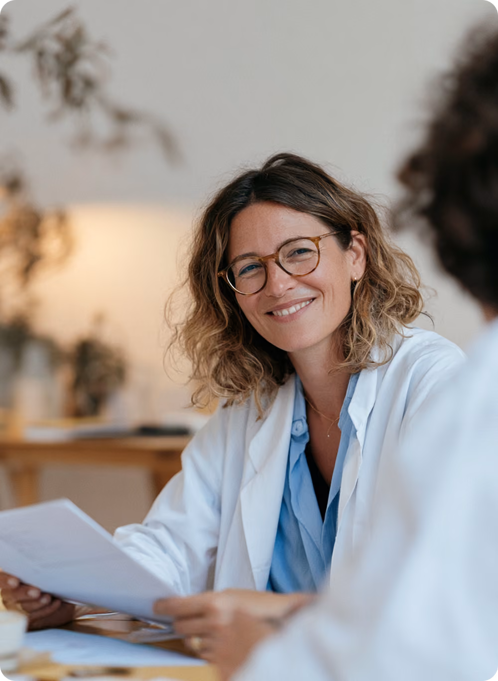 Médecin souriante portant une blouse blanche et des lunettes, tenant des documents, assise à une table avec une autre personne floue en avant-plan.