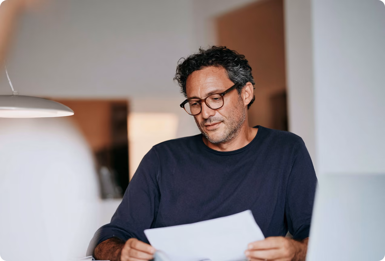 Homme portant des lunettes et un t-shirt bleu foncé, assis à une table et lisant un document papier.