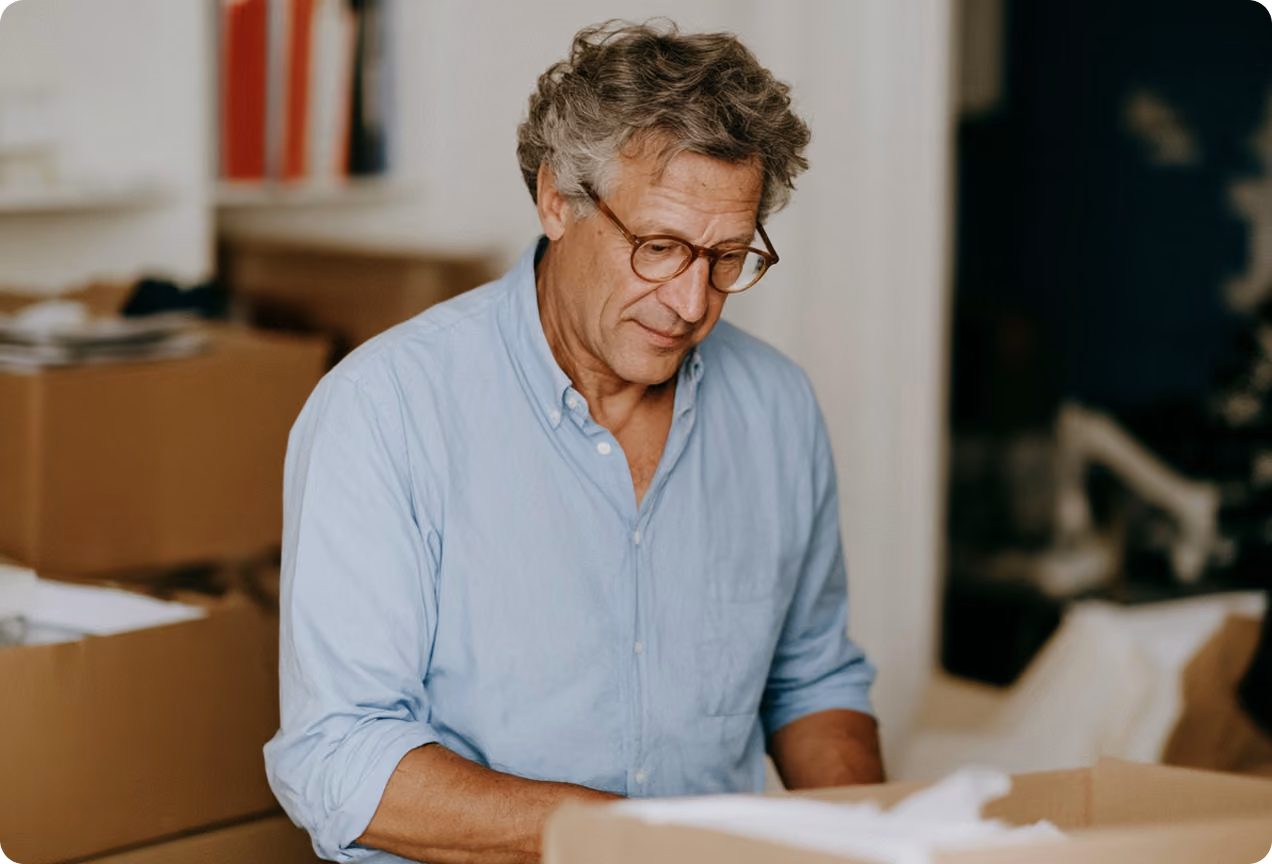 Homme âgé portant des lunettes et une chemise bleue claire, regardant attentivement des objets dans une boîte en carton dans une pièce avec des cartons empilés.