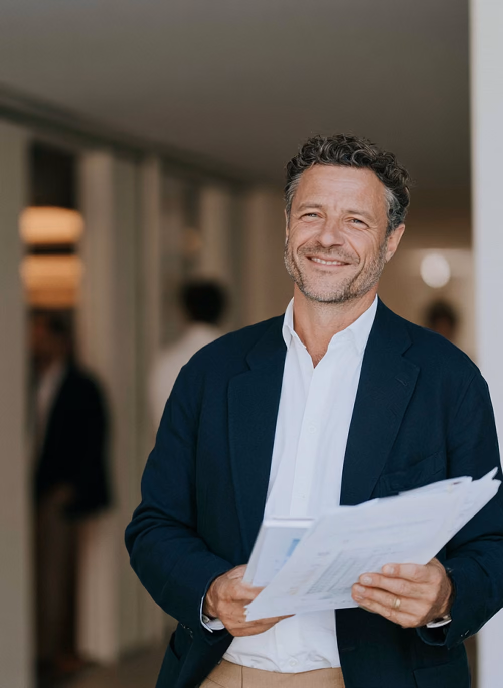 Homme d'affaires souriant portant une veste bleue marine et une chemise blanche, tenant des documents dans un couloir de bureau.