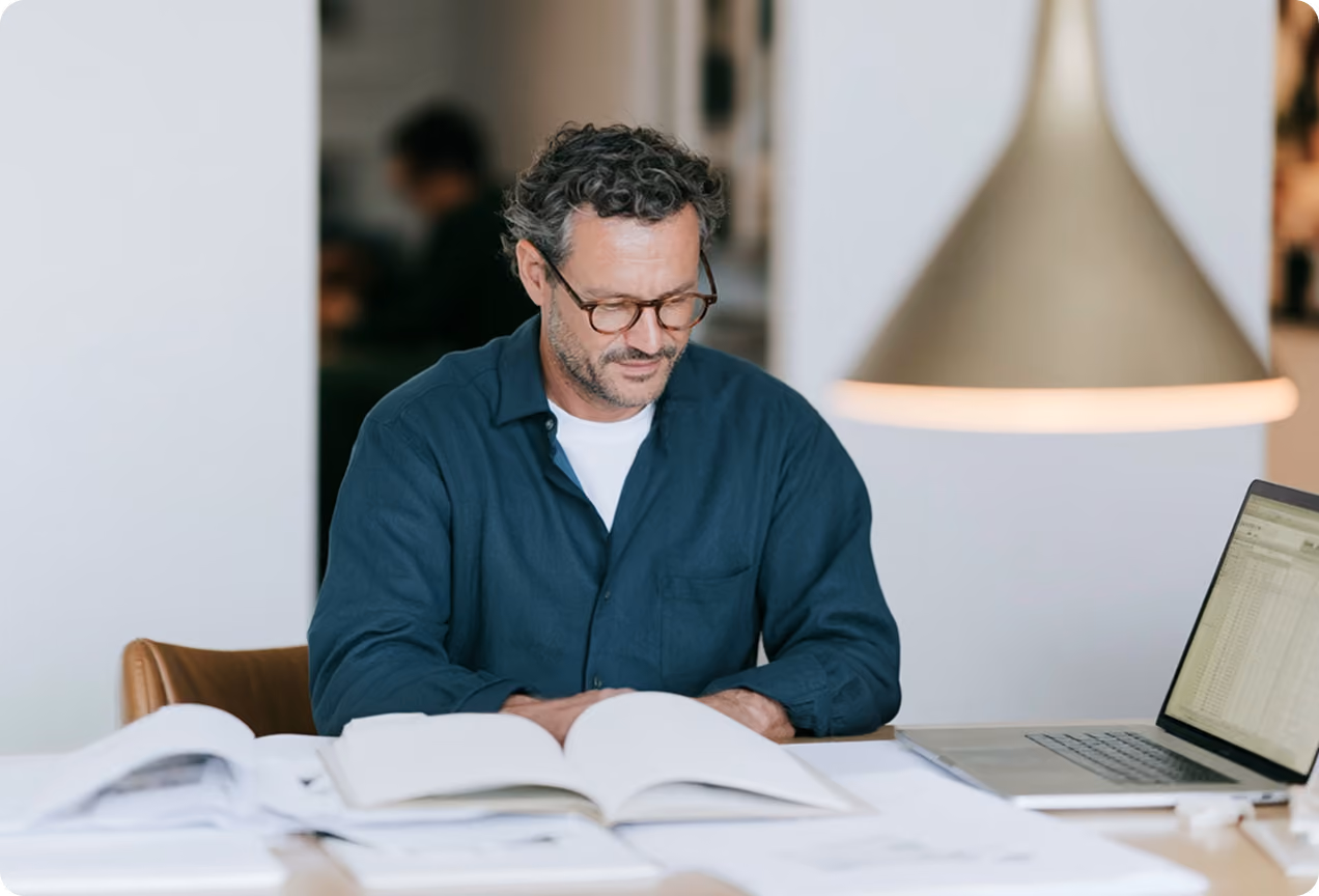 Homme portant des lunettes, assis à une table avec plusieurs documents ouverts et un ordinateur portable, travaillant dans un environnement intérieur lumineux.