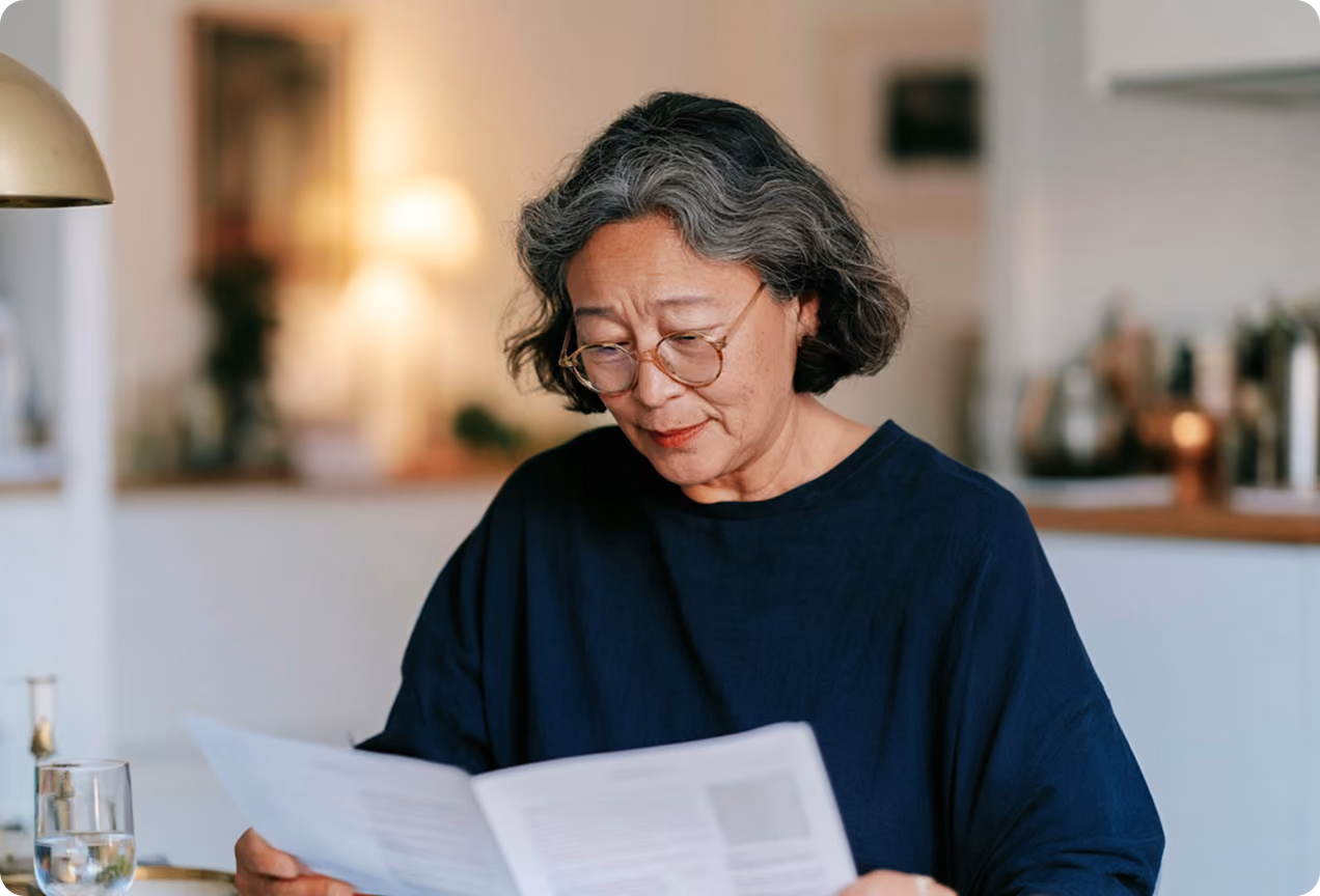 Une femme âgée aux cheveux gris et lunettes portant un pull bleu foncé lit attentivement un document dans une pièce éclairée.