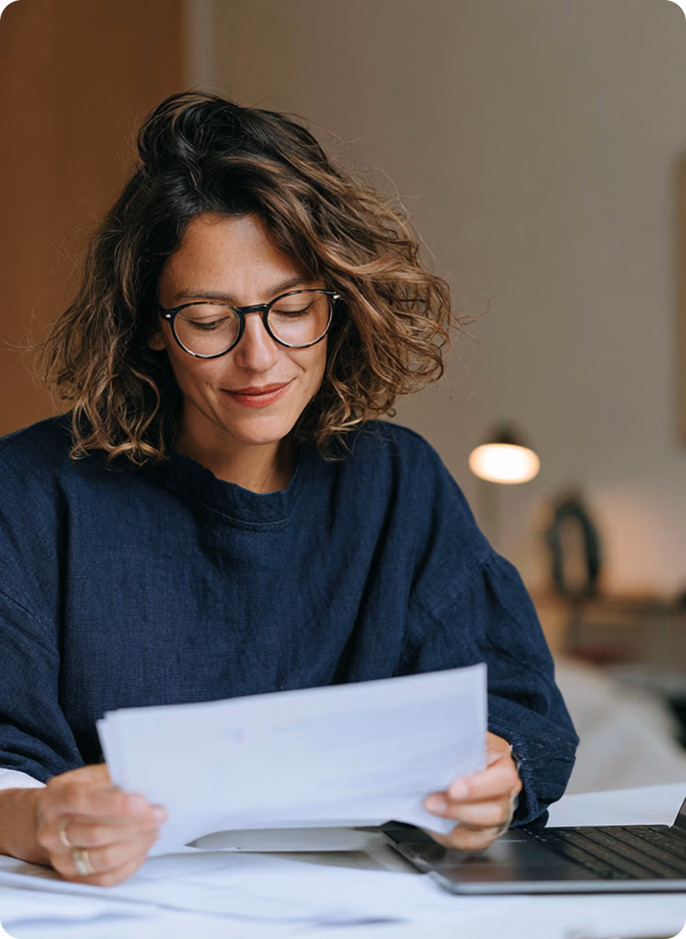 Femme souriante avec des lunettes, cheveux bouclés, en chemise bleue sombre, lisant un document papier près d'un ordinateur portable dans un cadre intérieur.