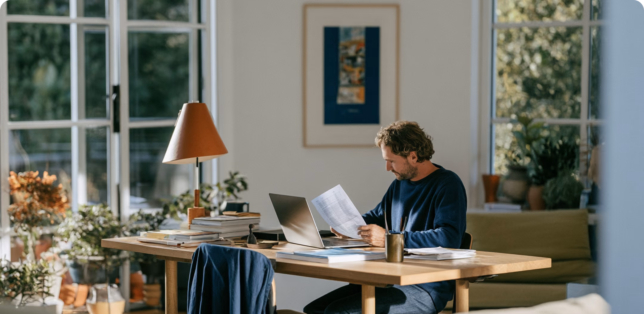Homme assis à une grande table en bois dans un espace lumineux avec de grandes fenêtres, travaillant sur un ordinateur portable et consultant un document papier, entouré de livres et de quelques plantes d'intérieur.