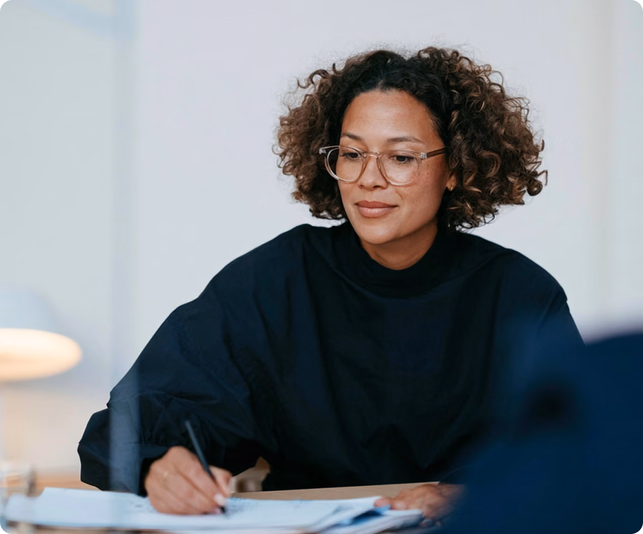 Une femme à cheveux bouclés portant des lunettes et un pull noir écrit à la main sur des documents posés sur une table dans un environnement intérieur clair.