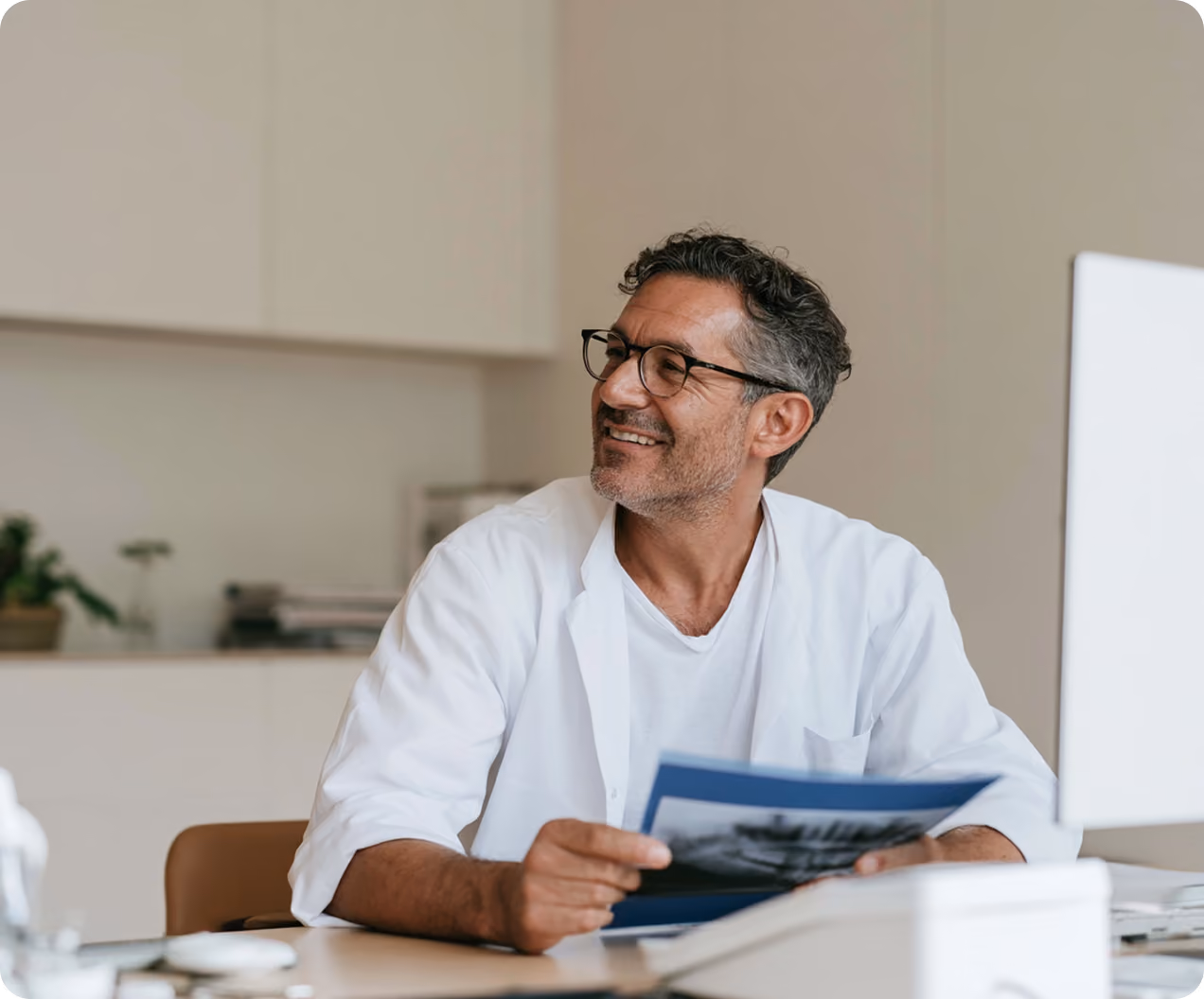 Homme souriant portant une blouse blanche et des lunettes, tenant un document assis à un bureau devant un ordinateur dans un bureau moderne.