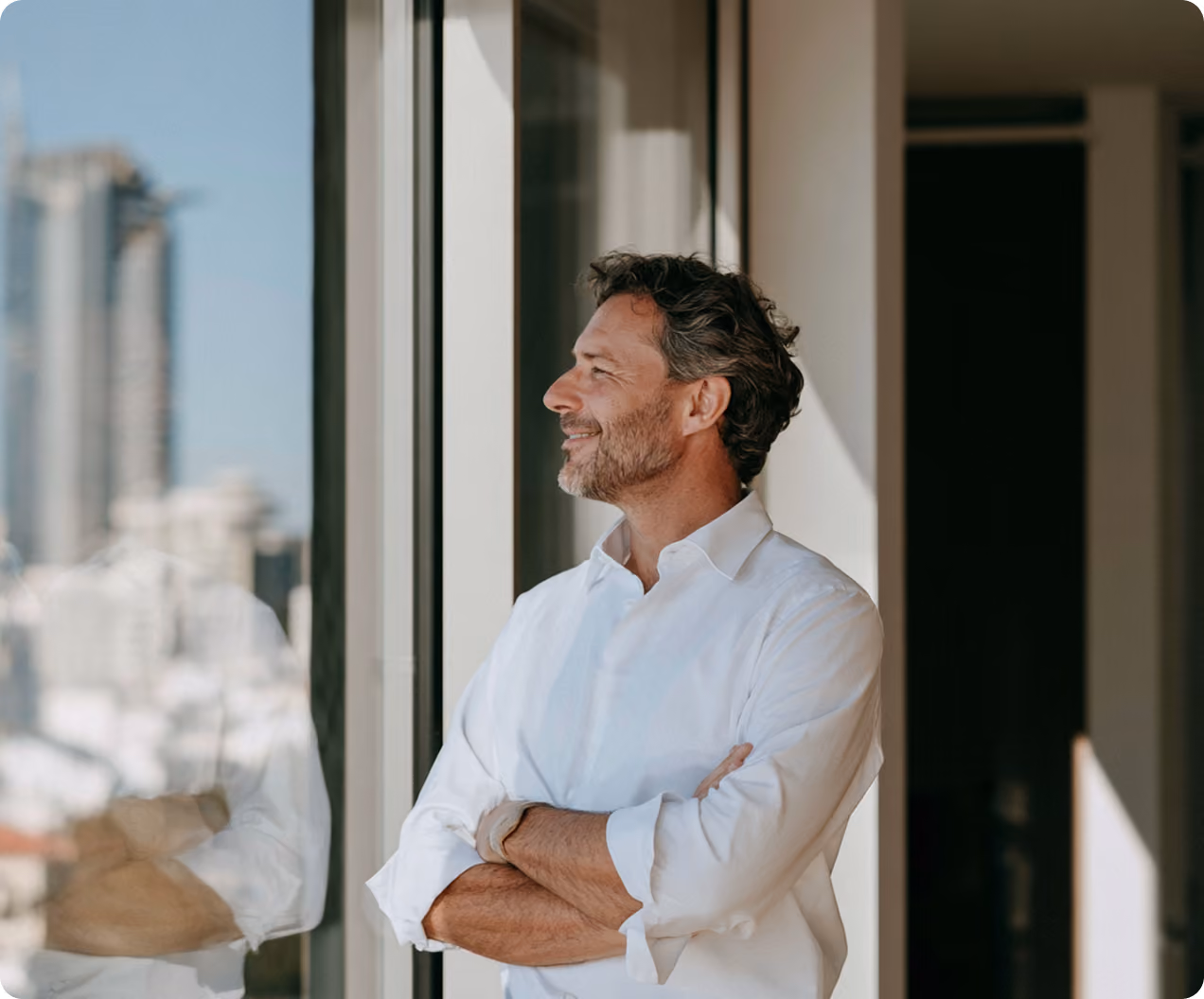 Homme souriant aux cheveux gris et barbe portant une chemise blanche, les bras croisés, regardant par une grande fenêtre avec vue urbaine floue.
