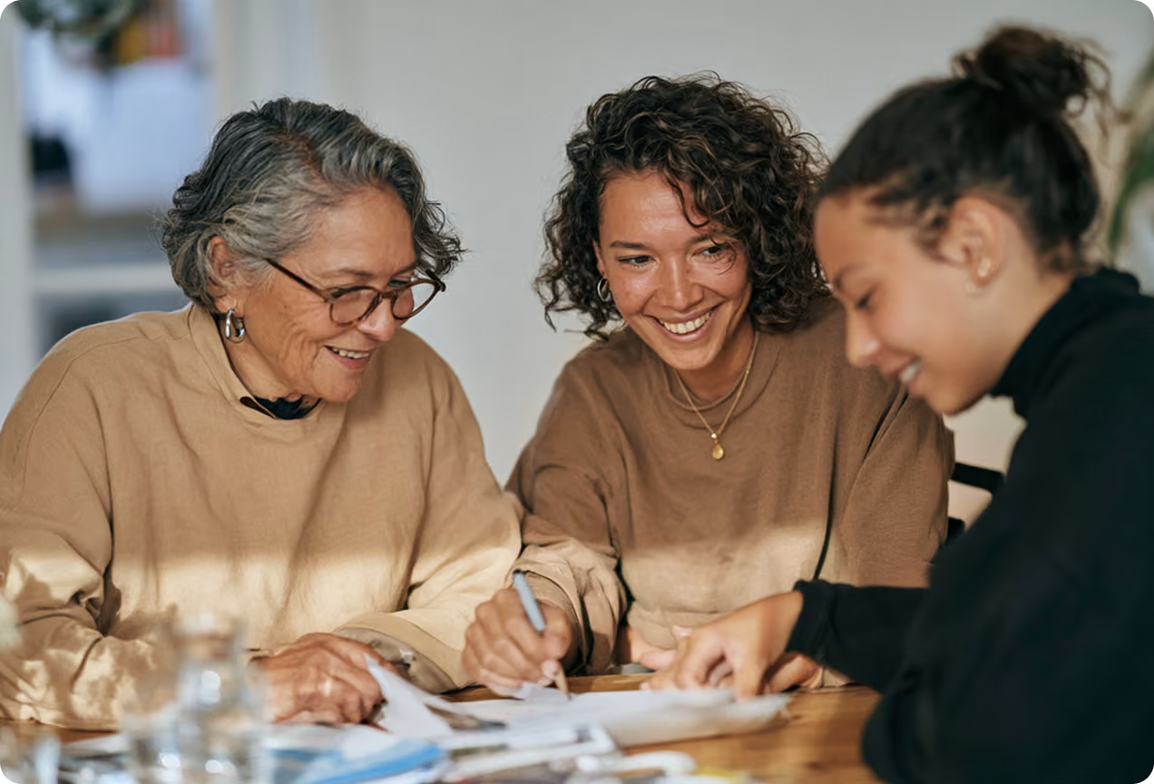 Trois femmes de différentes générations souriantes, assises ensemble à une table, collaborant et écrivant sur des documents.
