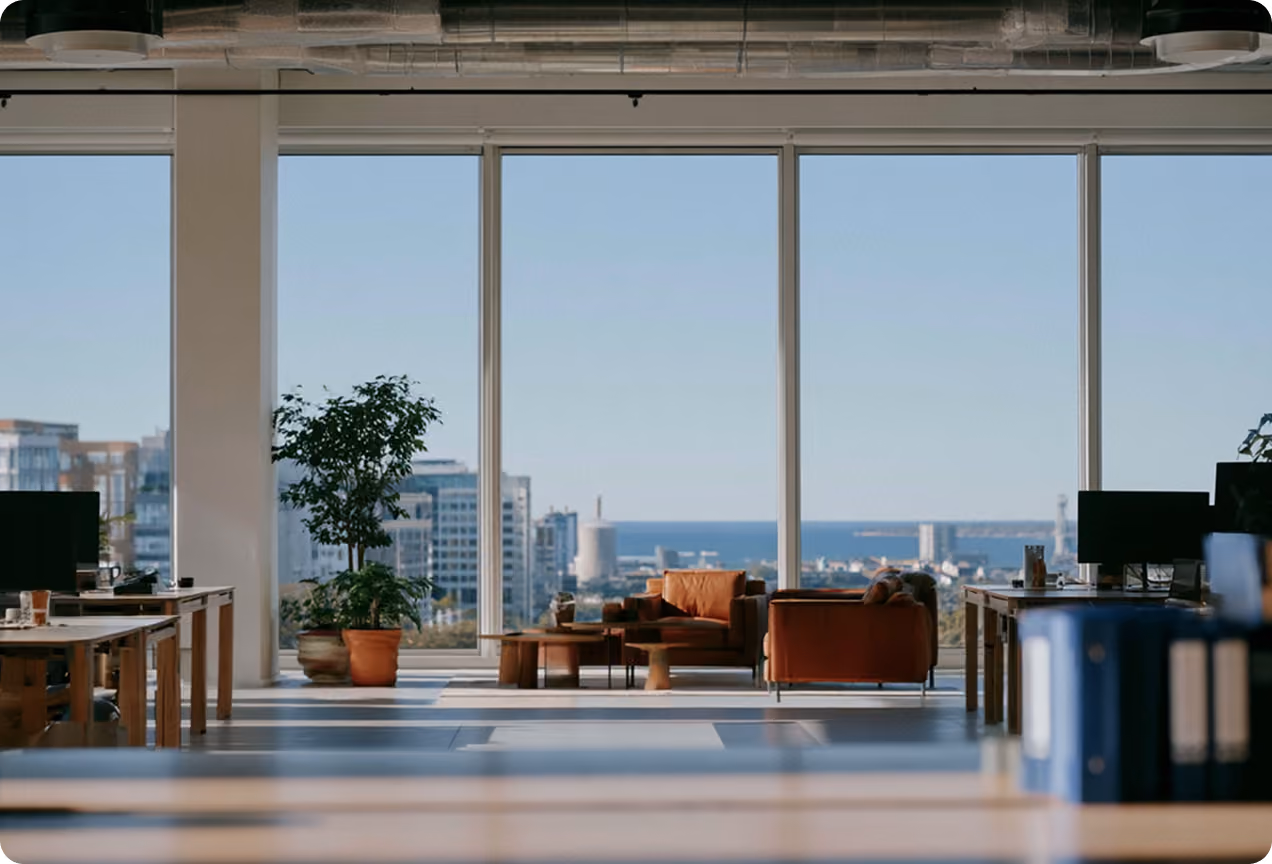 Bureau moderne avec grandes fenêtres offrant une vue sur la ville, des canapés en cuir marron et des plantes en pot.