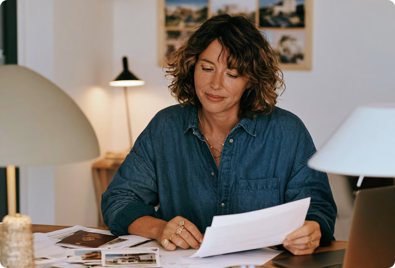 Femme aux cheveux bouclés portant une chemise en jean, assise à une table en bois, examinant des documents papier avec un ordinateur portable à côté d'elle dans une pièce lumineuse avec deux lampes.