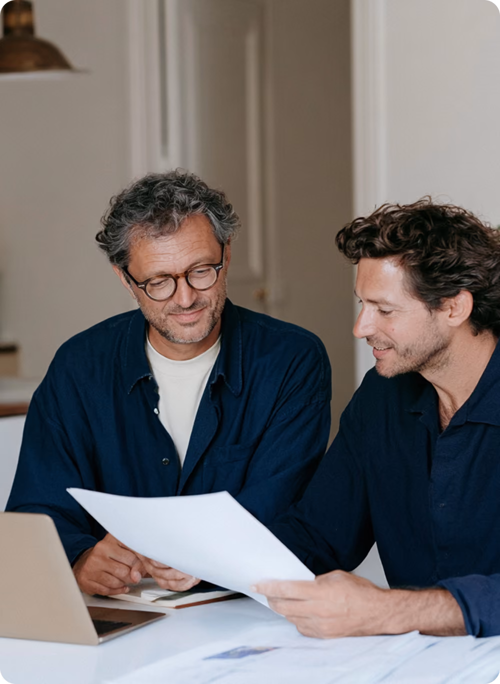 Deux hommes souriants discutant ensemble autour d'une table avec un ordinateur portable ouvert, l'un tenant une feuille de papier tandis que l'autre regarde le document.