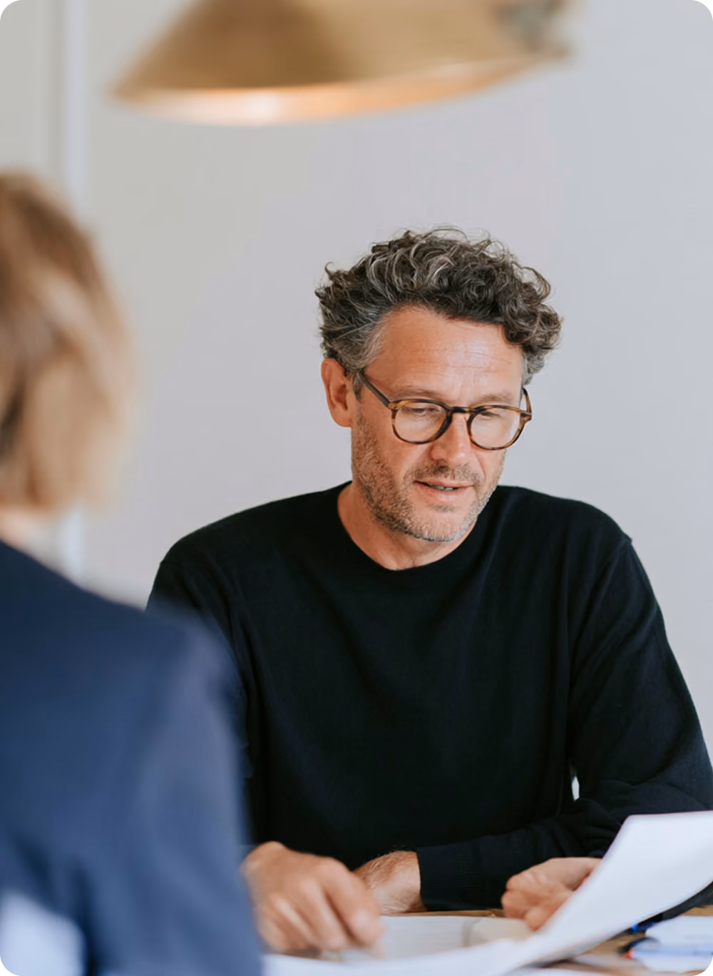 Un homme aux cheveux bouclés et gris portant des lunettes et un pull noir lit attentivement un document, tandis que la personne en vis-à-vis est floue au premier plan.