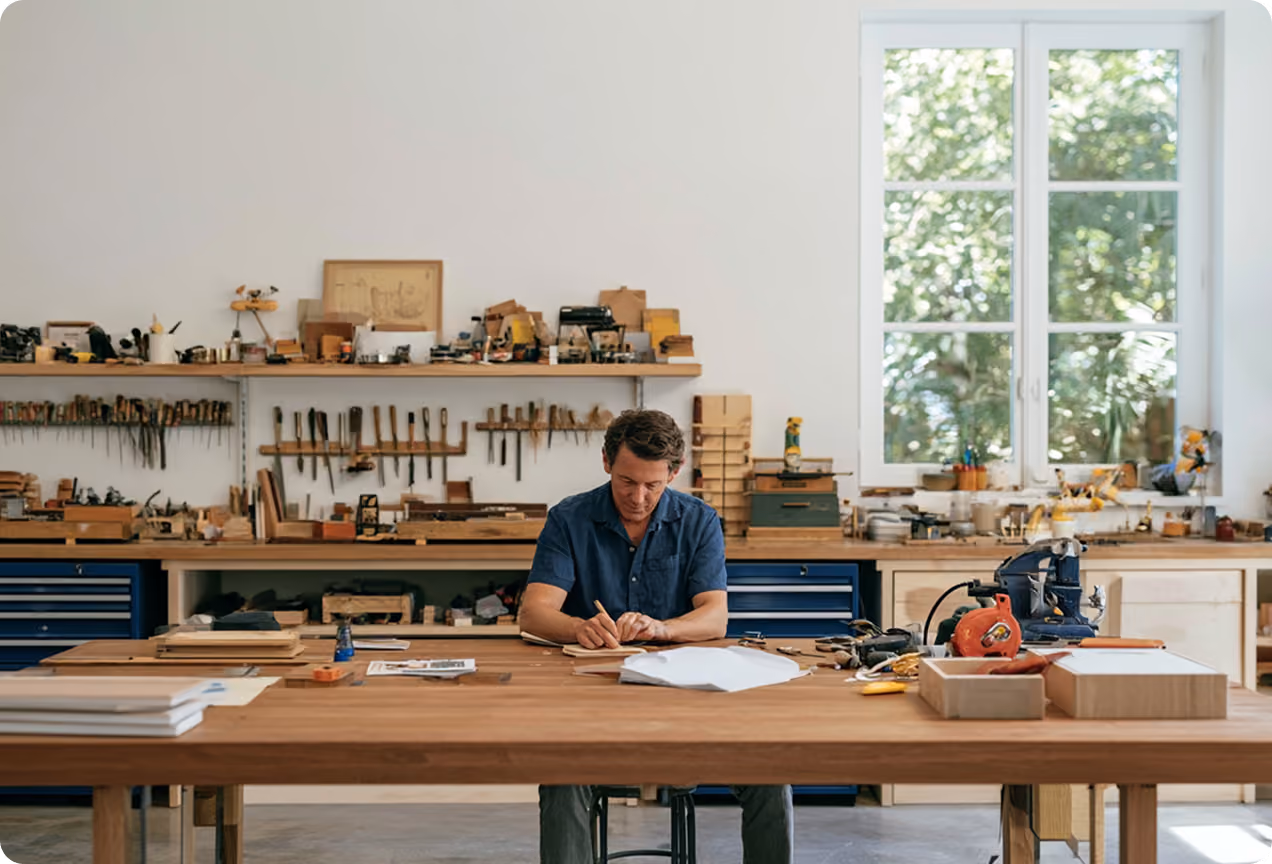 Homme travaillant sur un projet de menuiserie dans un atelier lumineux avec des outils accrochés au mur et une grande fenêtre laissant entrer la lumière naturelle.