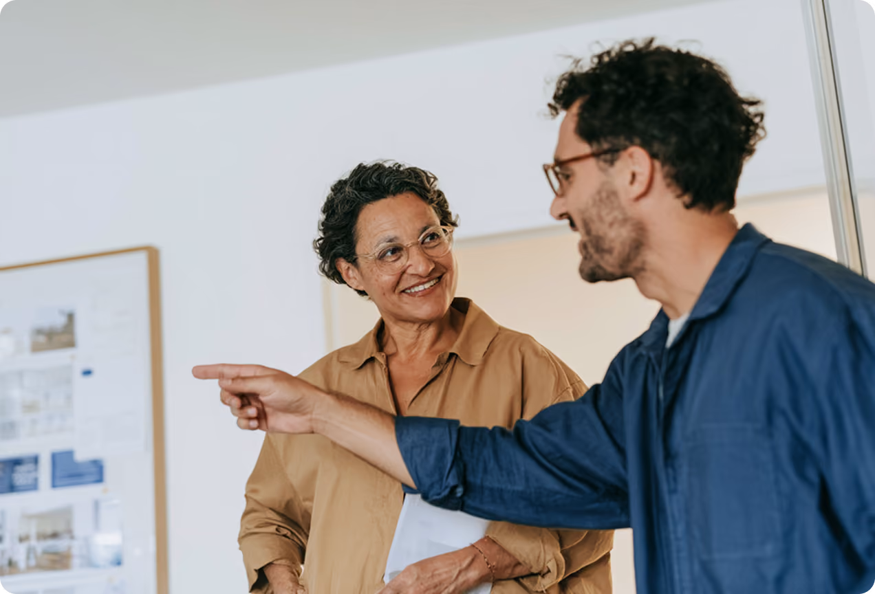 Un homme avec des lunettes et une barbe portant une combinaison bleue pointe du doigt pendant qu'une femme souriante avec des lunettes et une chemise beige le regarde dans une pièce claire avec des documents affichés sur un mur.