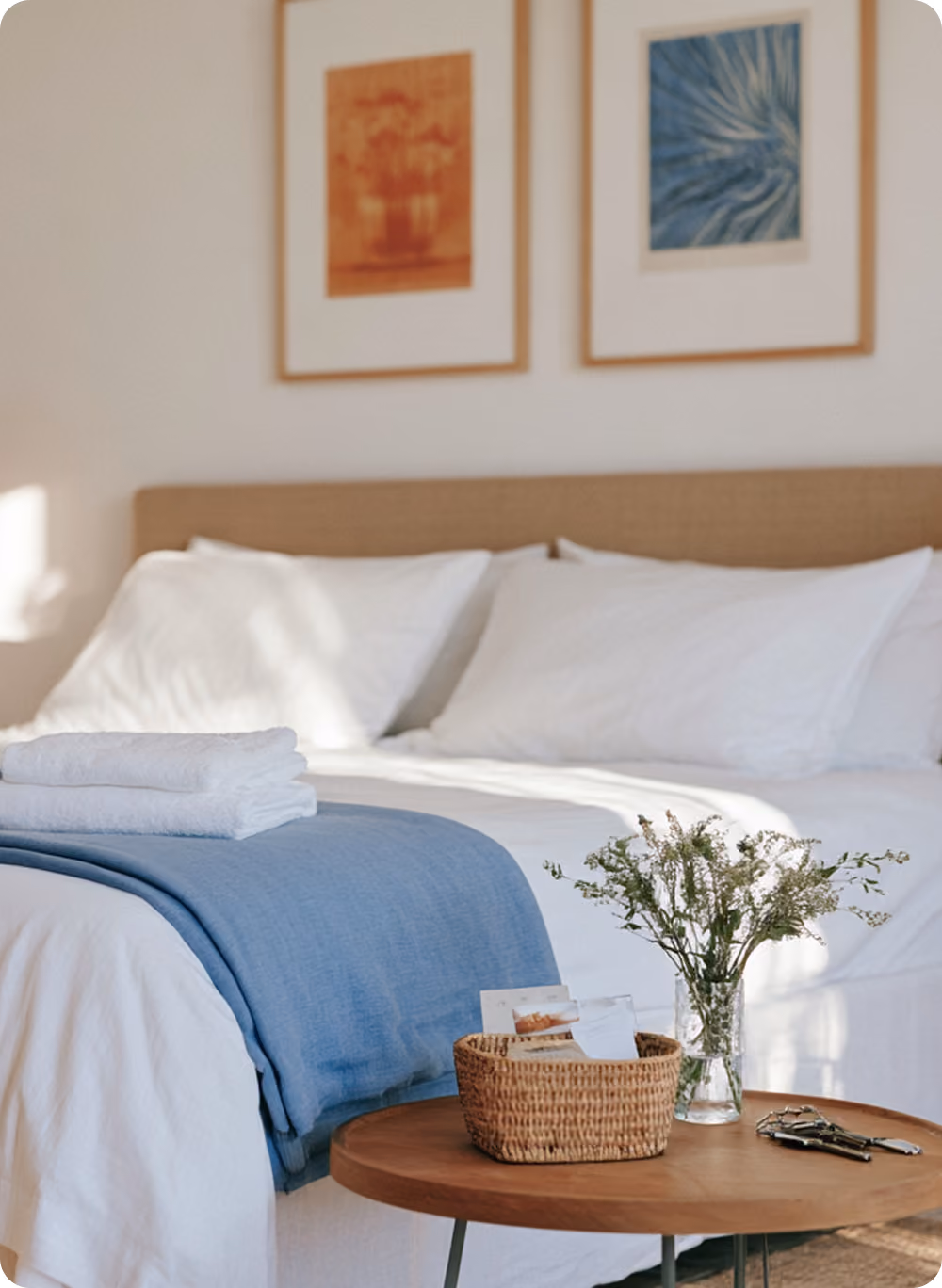 Chambre claire avec grand lit fait aux draps blancs et couverture bleue, serviettes pliées dessus, tableau et vase de fleurs blanches sur table en bois ronde devant.