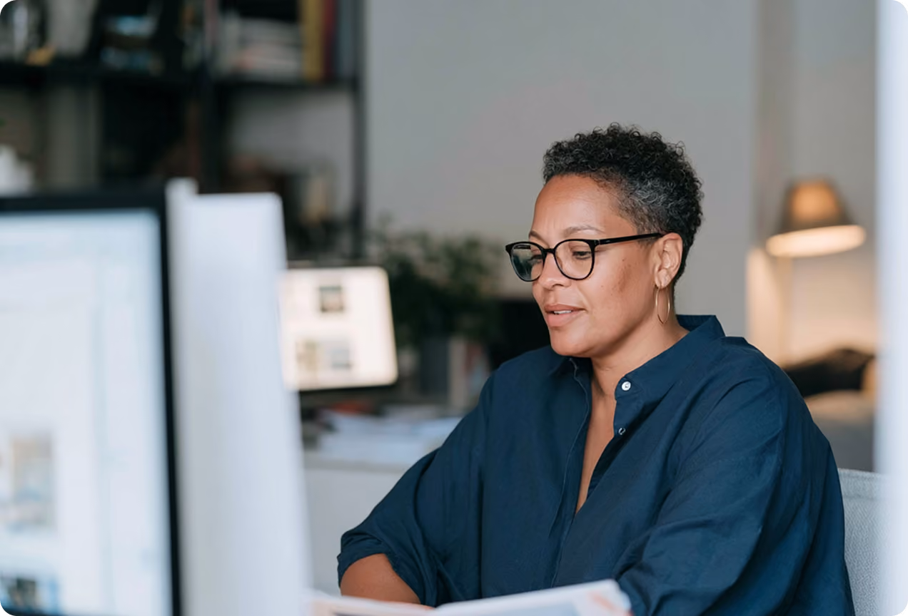 Une femme portant des lunettes et une chemise bleu foncé est assise devant un ordinateur dans un bureau, concentrée sur son travail.