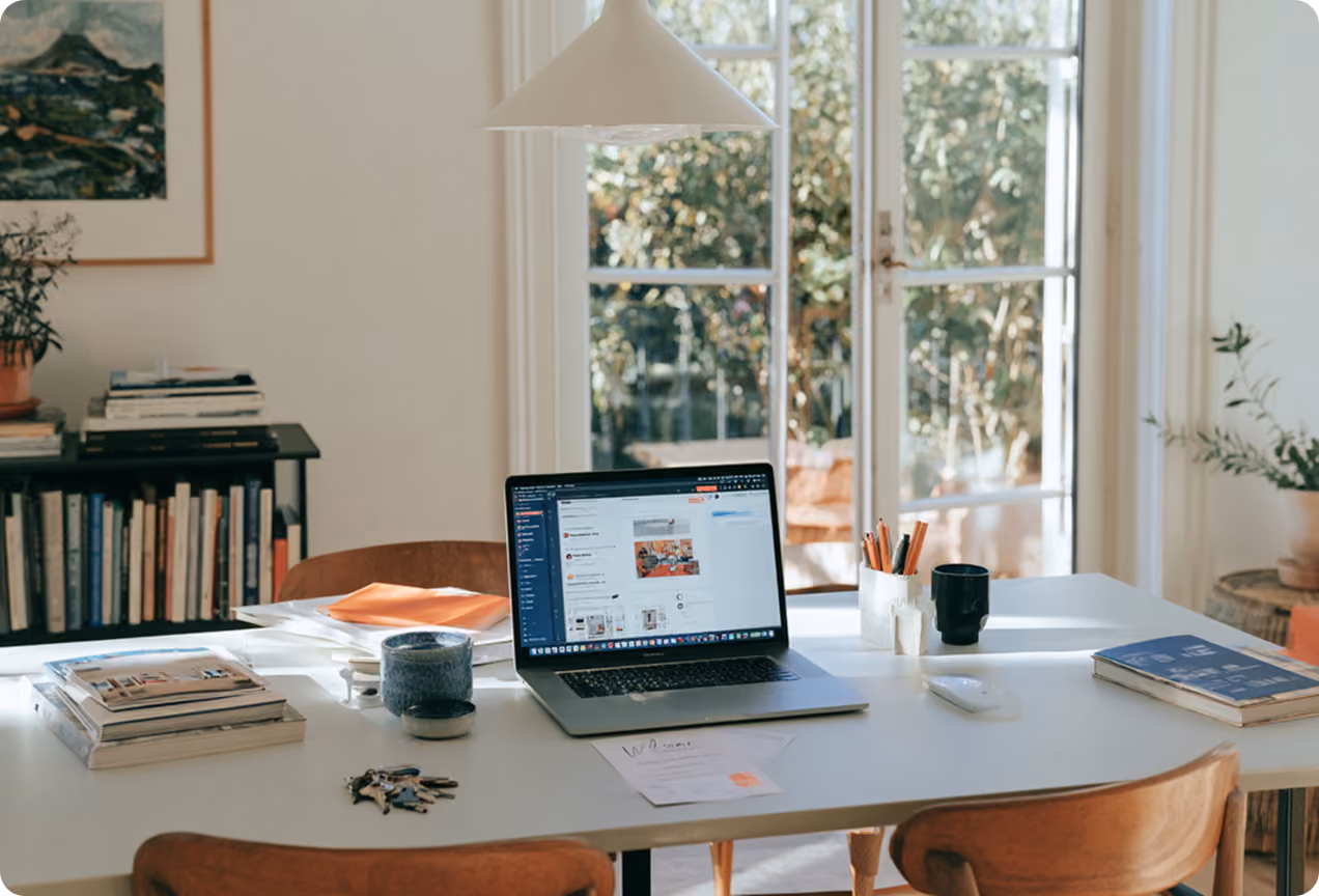Un bureau blanc près de grandes portes vitrées avec vue sur un jardin, avec un ordinateur portable ouvert, des livres, des stylos, une tasse et des clés posés dessus.
