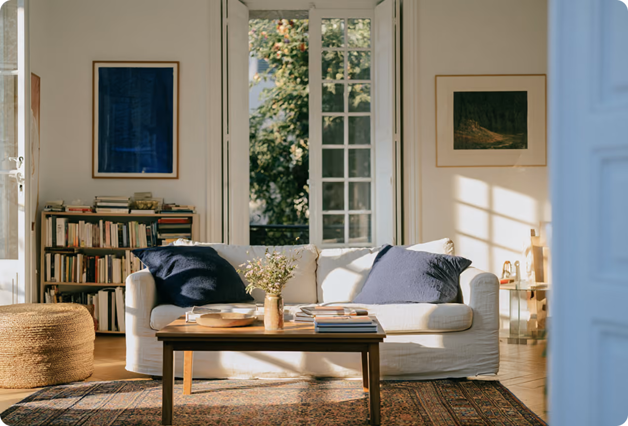 Salon lumineux avec canapé blanc et coussins bleus foncés, table basse en bois avec livres et vase de fleurs, bibliothèque remplie de livres à gauche, grandes fenêtres ouvertes laissant entrer la lumière naturelle.