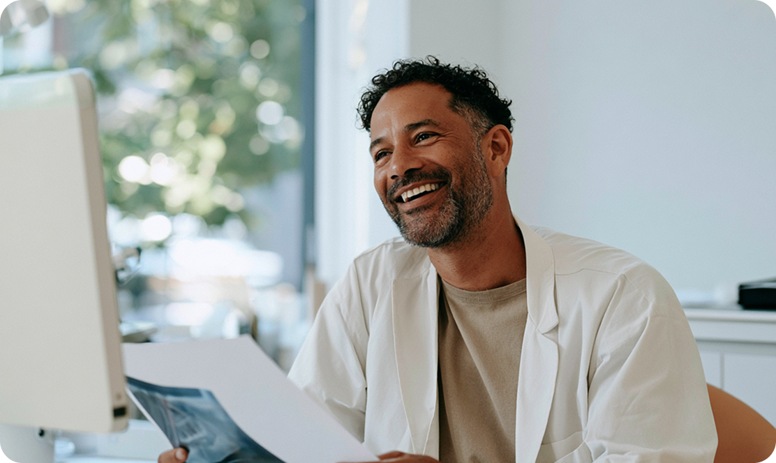 Un homme souriant portant une blouse blanche regarde un écran d'ordinateur en tenant des documents dans un bureau lumineux.