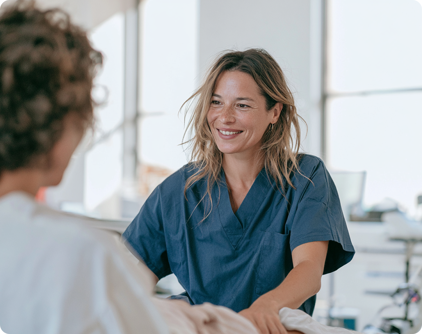 Femme en blouse médicale bleue souriant en parlant avec une autre personne dans un environnement clair et professionnel.