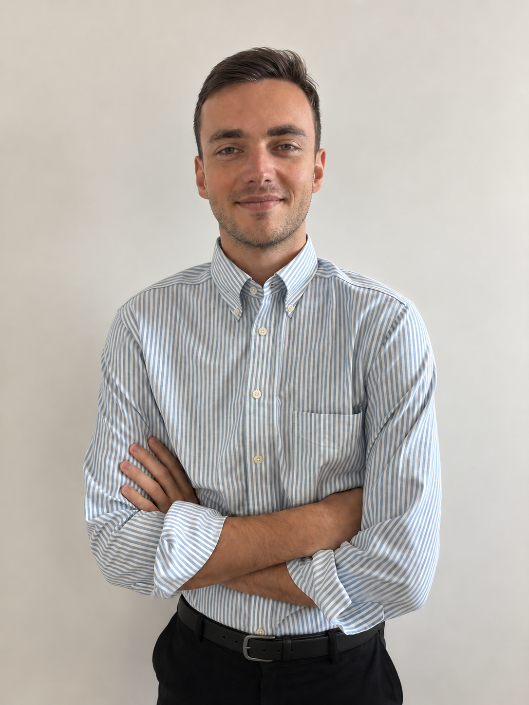 Homme jeune debout, bras croisés, portant une chemise à rayures bleu clair et blanche et un pantalon noir, devant un fond uni beige clair.