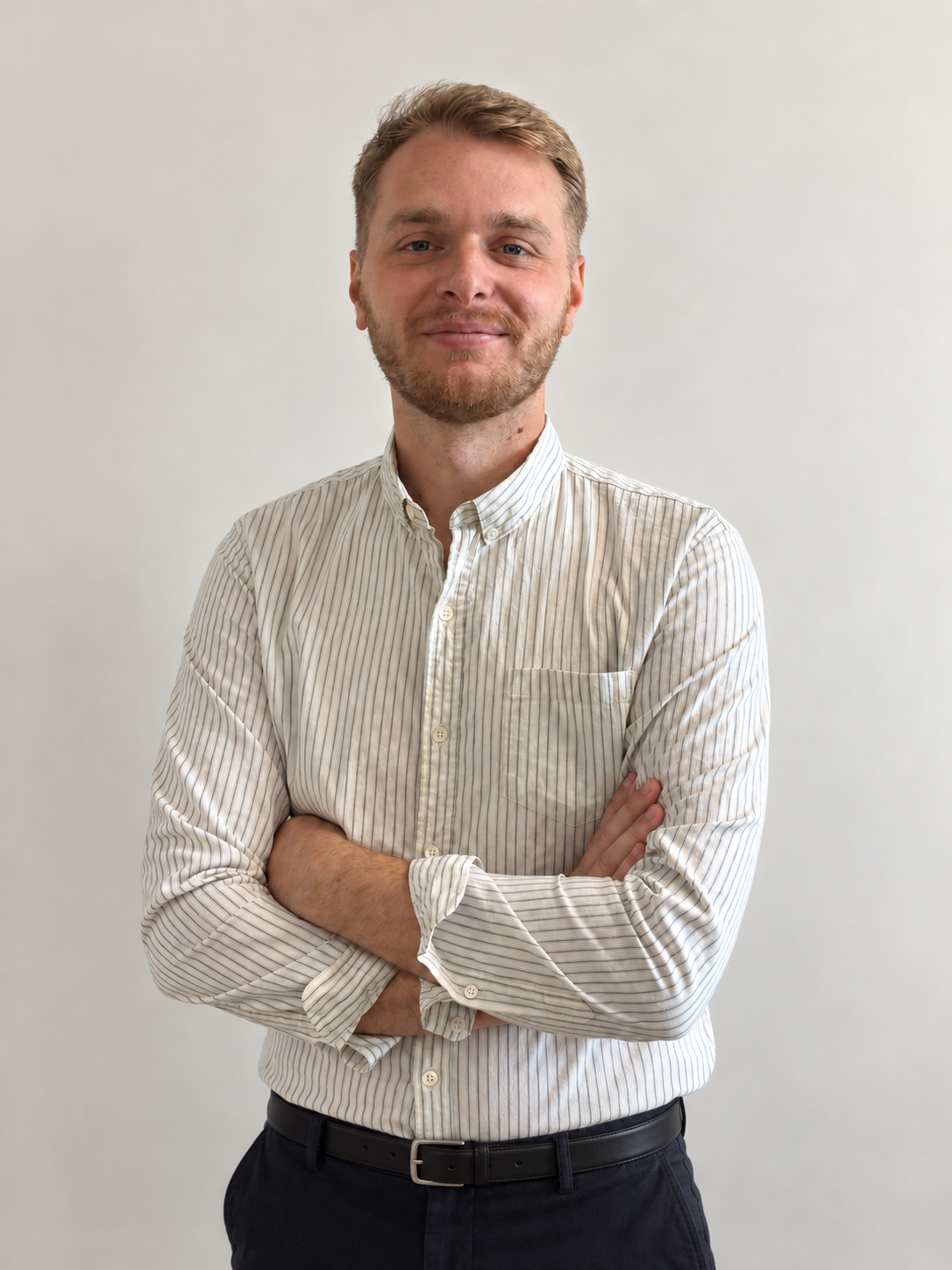 Homme souriant aux cheveux blonds courts et barbe légère, portant une chemise blanche à fines rayures noires et un pantalon noir, bras croisés, sur fond gris clair.