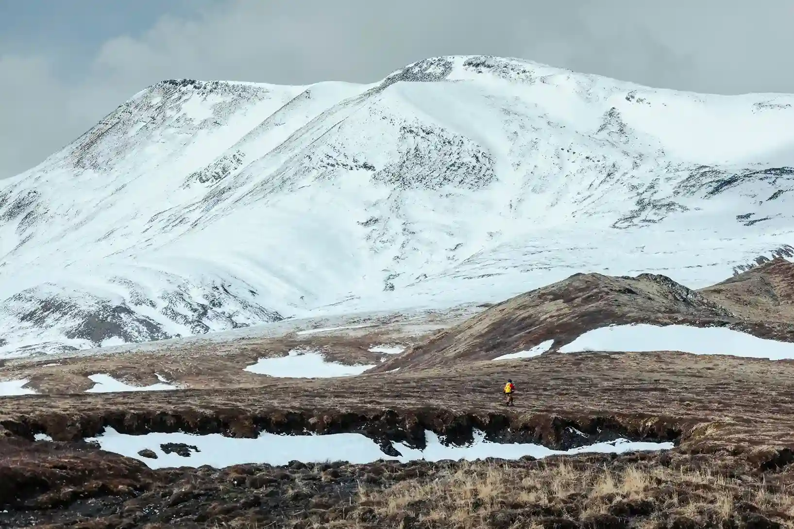 A single figure crossing tundra below a snow-covered mountain ridge in the Arctic