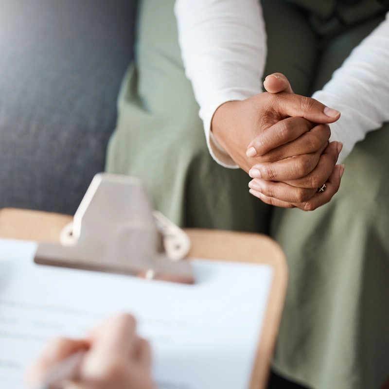 Close-up of two people during a consultation: one person with clasped hands resting on their lap, the other holding a pen and clipboard with a blurred document.