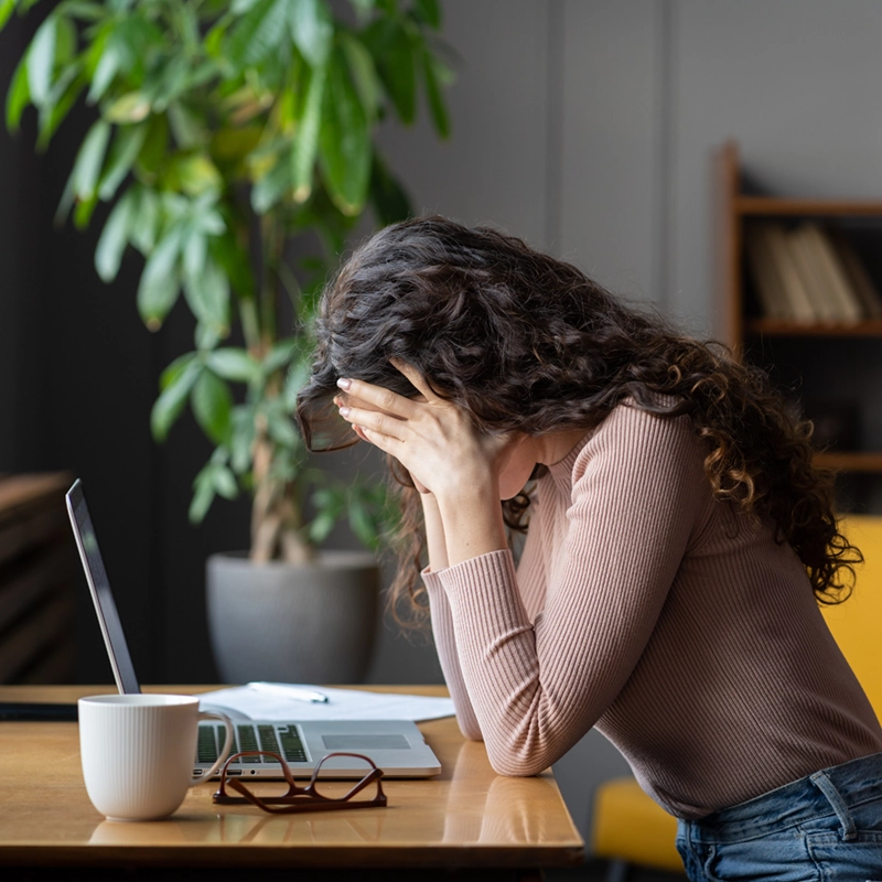 A woman with curly hair sits at a table with her face in her hands looking stressed, next to a laptop, glasses, a cup, and papers, with a large potted plant and bookshelf in the background.