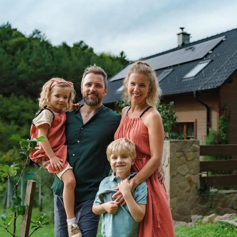Smiling family of four posing outdoors in front of a house with solar panels on the roof; the father with a beard holds a young girl in a pink dress, while the mother in a matching dress stands beside a blond boy in a light blue shirt.