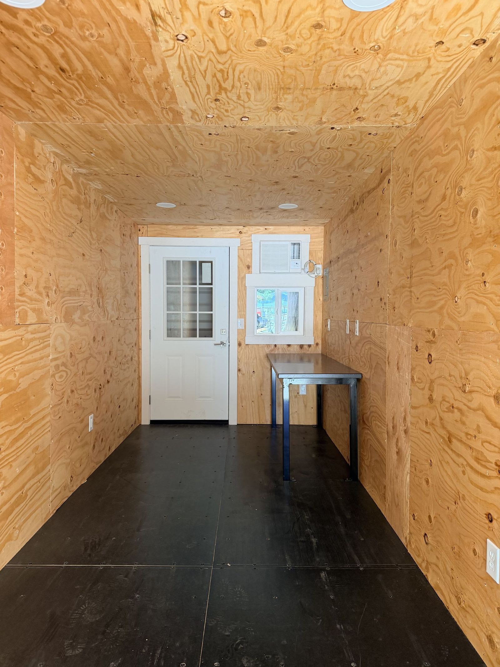 Plywood-lined container office interior with a personnel door, window, air conditioner, and desk