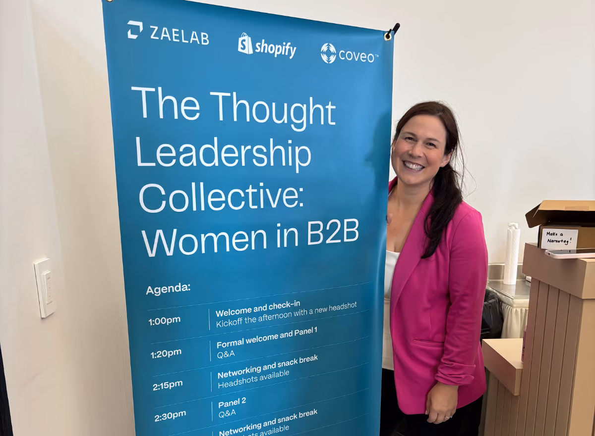 A smiling attendee poses next to the event agenda banner for the Women in B2B Thought Leadership Collective, sponsored by Zaelab, Shopify, and Coveo.