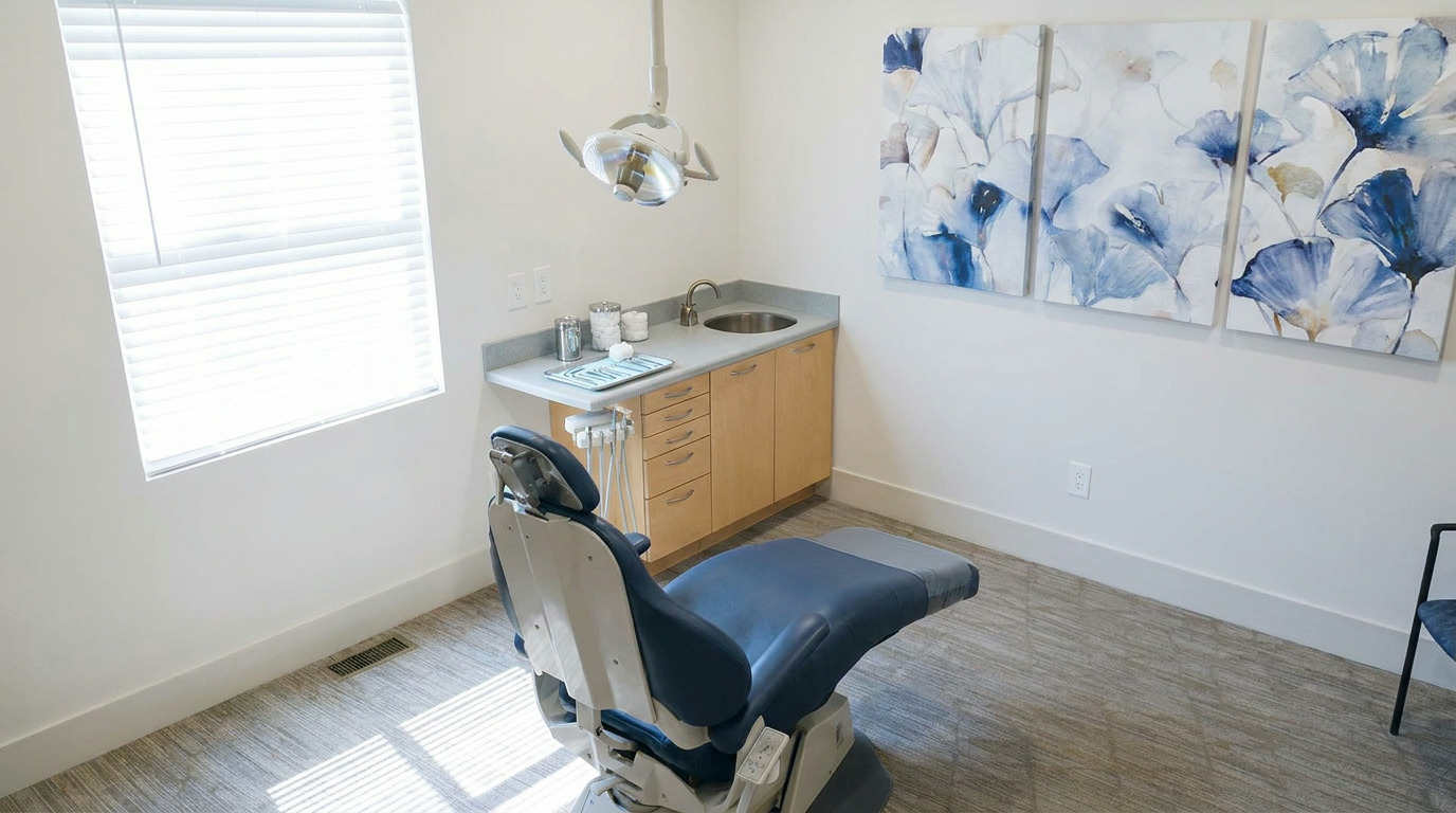 A modern dental operatory features an empty blue and gray dental chair, a window with blinds, a counter with a sink and instruments, a dental light, three blue floral artworks on the wall, and textured carpeting.