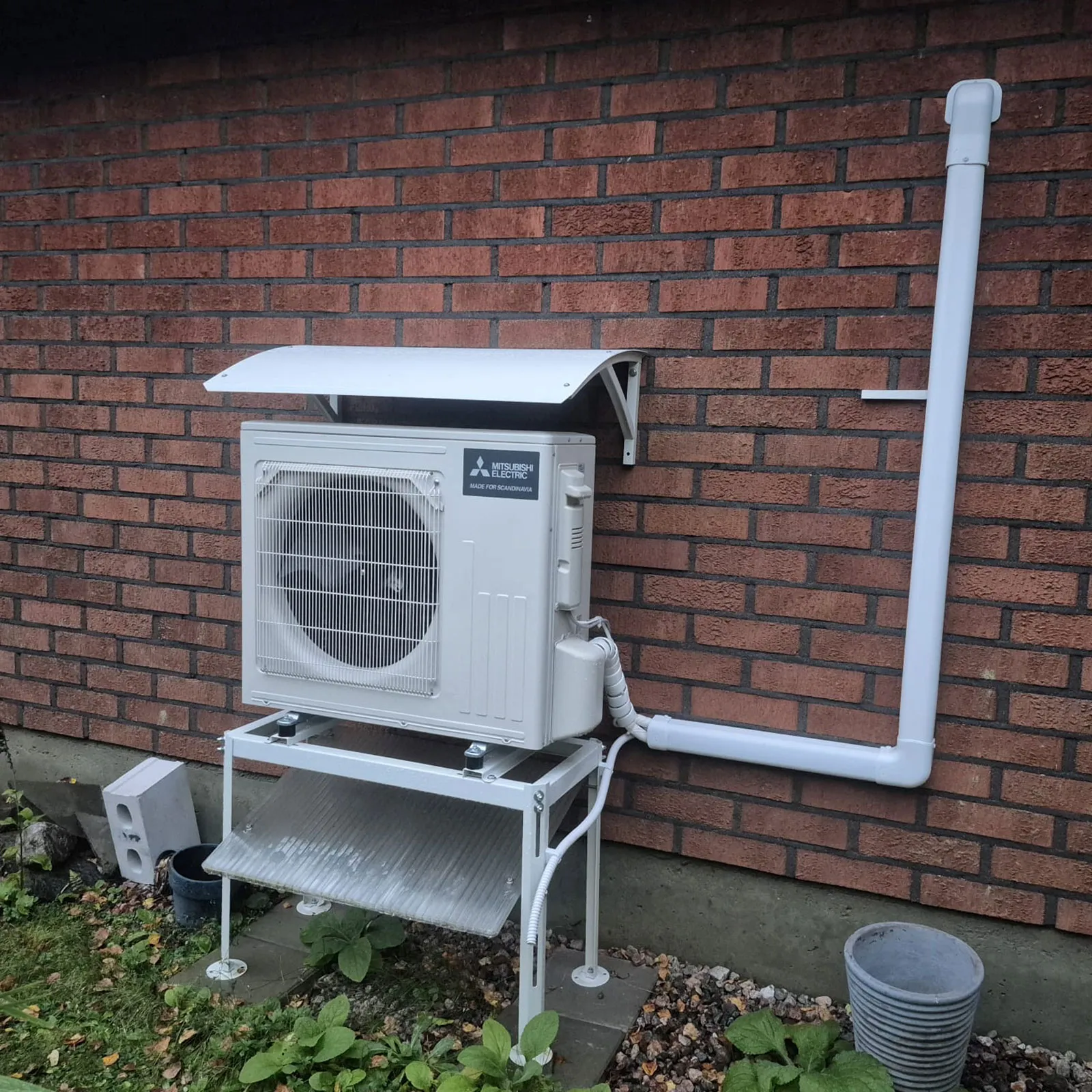 Mitsubishi Electric -ilmalämpöpumpun ulkoyksikkö jalustalla ja suojakatteen alla tiiliseinän vieressä